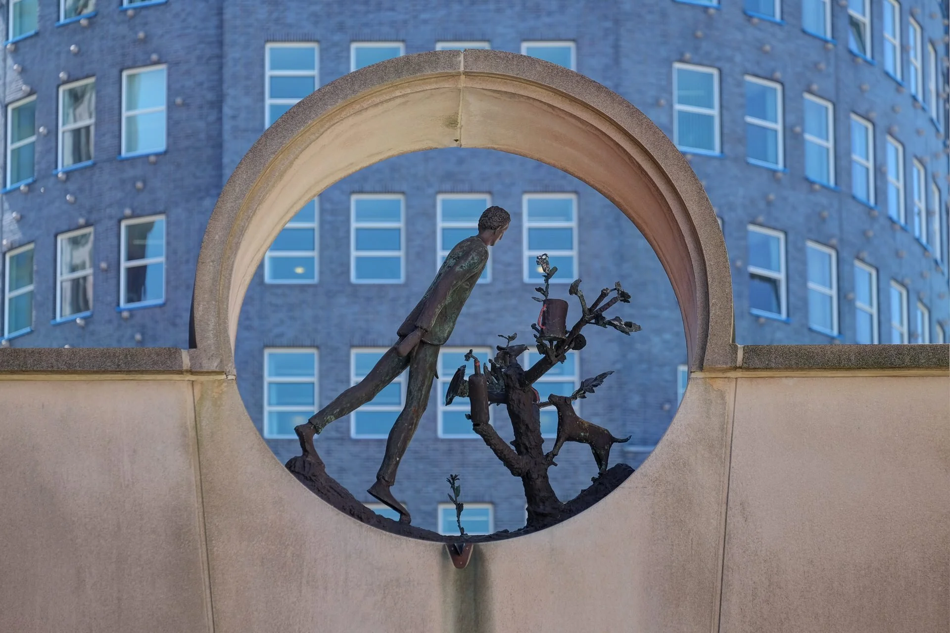 A bronze sculpture of a man walking up a ladder, surrounded by tree branches, beneath a stone archway with a modern multi-story building with numerous windows in the background.