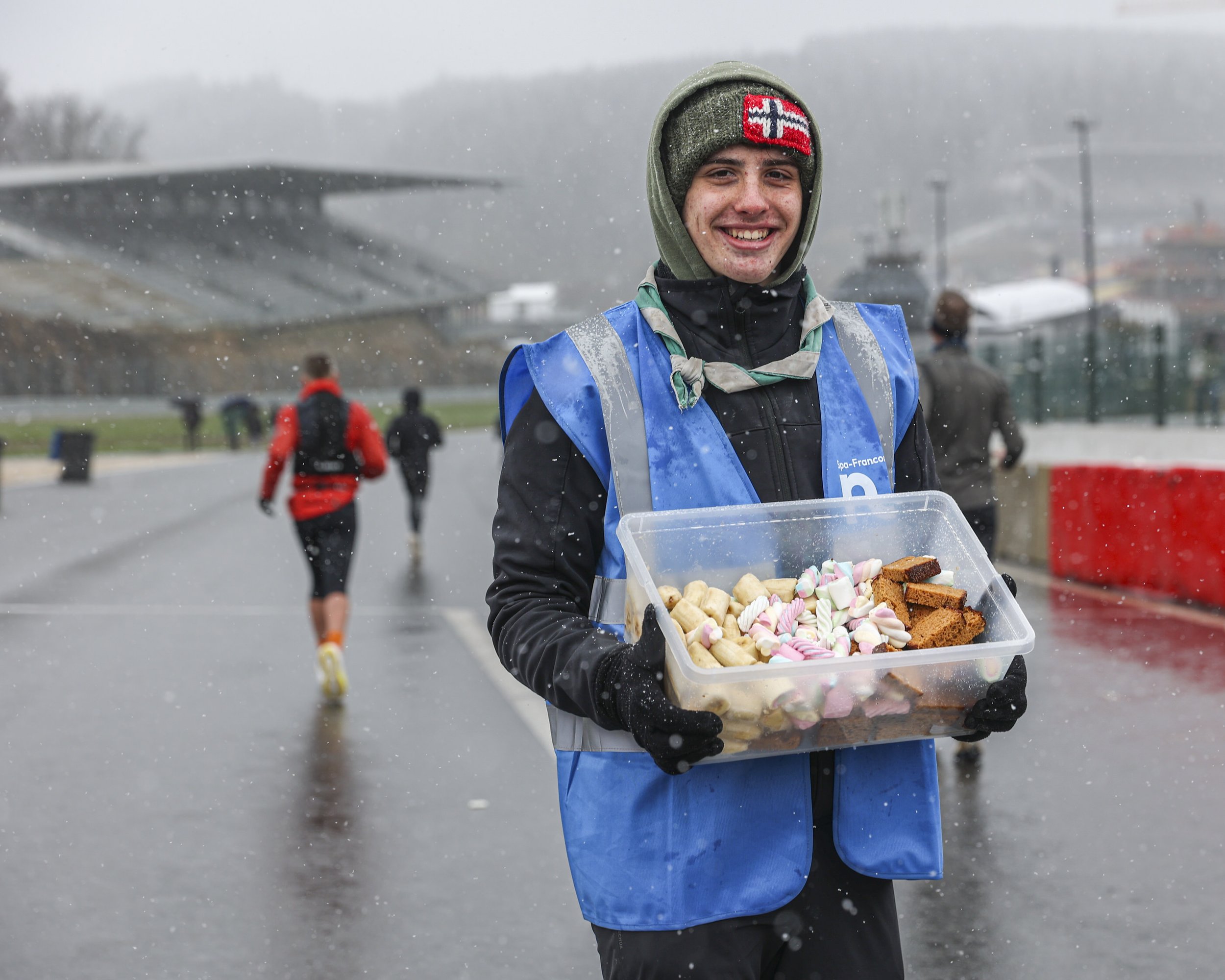 A smiling person wearing a winter hat and jacket, holding a container of assorted marshmallows and baked treats, standing in a snowy outdoor environment with other people in the background.