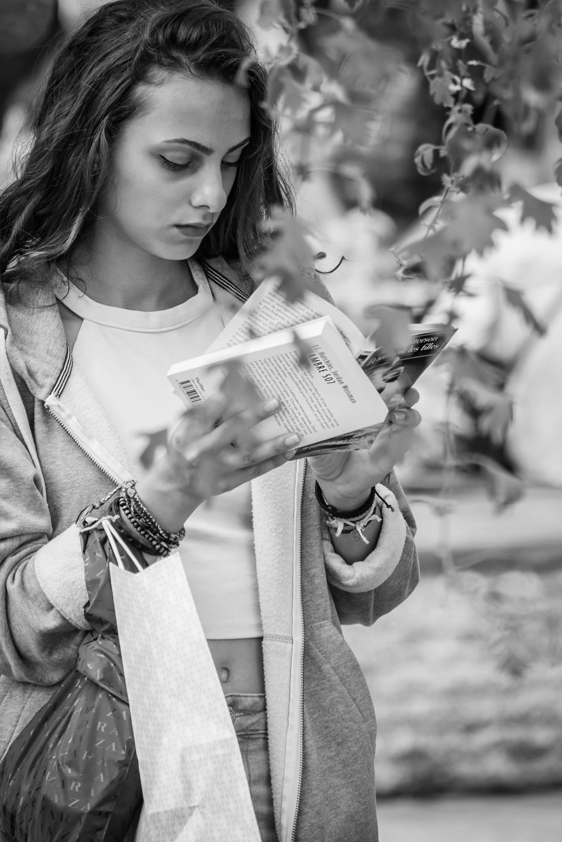 A young woman with long hair, wearing a zip-up hoodie, is reading a book or a pamphlet while standing outdoors among foliage.