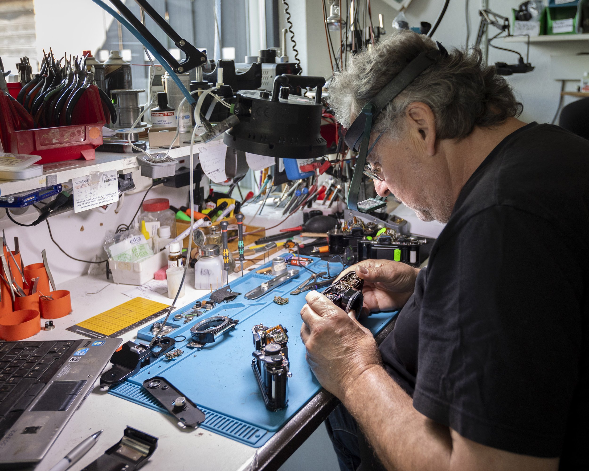 An elderly man working on electronic devices and circuit boards at a cluttered workbench in a workshop. He is wearing protective glasses and headphones, focused on repairing a device.