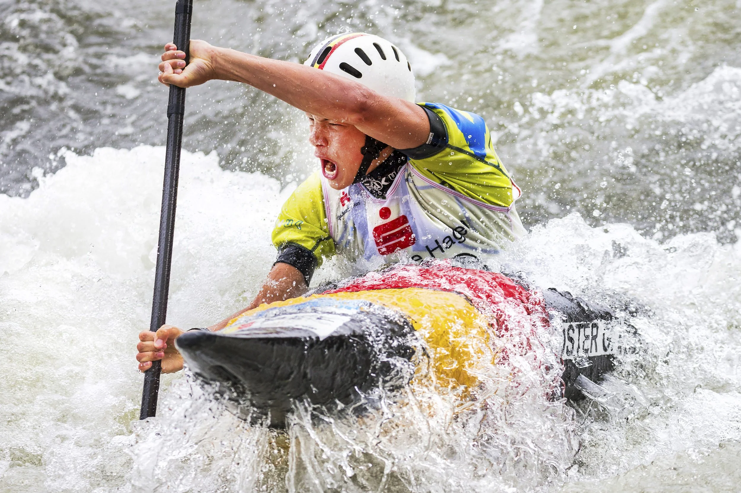 A person wearing a white helmet and a yellow and blue life vest is rafting in rough water, leaning forward with an intense expression and holding a paddle. Water splashes around as they navigate.