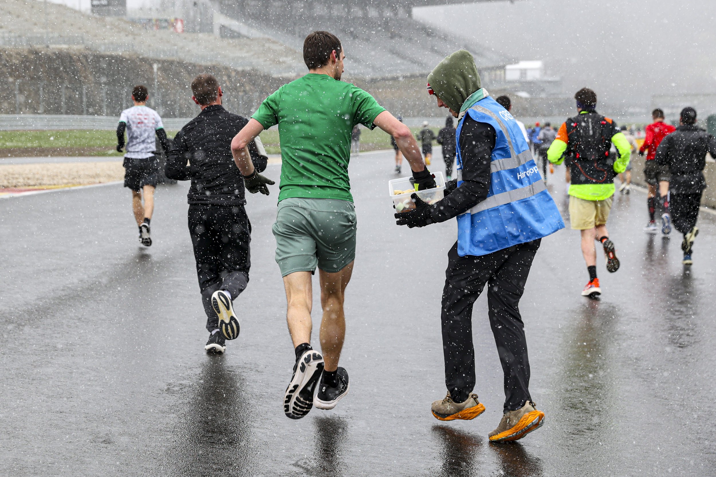 A group of runners participating in a race during rainy weather, with one runner in green shirt and another in camouflage shoes in the foreground, and others in colorful attire further back.