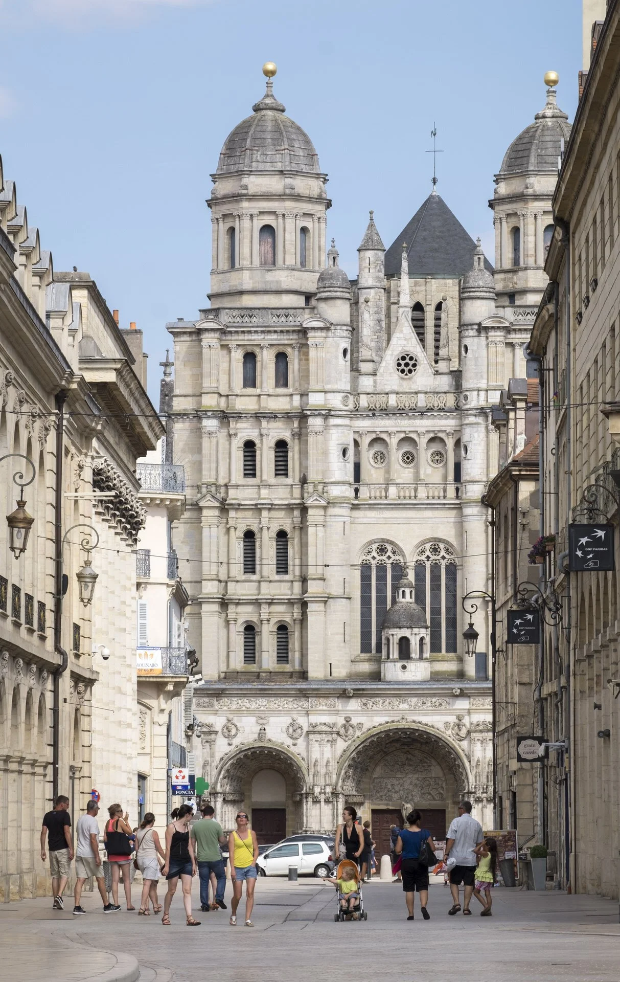 A busy European city square with people walking, including families and individuals, in front of a large historic cathedral with intricate stonework and twin towers topped with golden spheres.