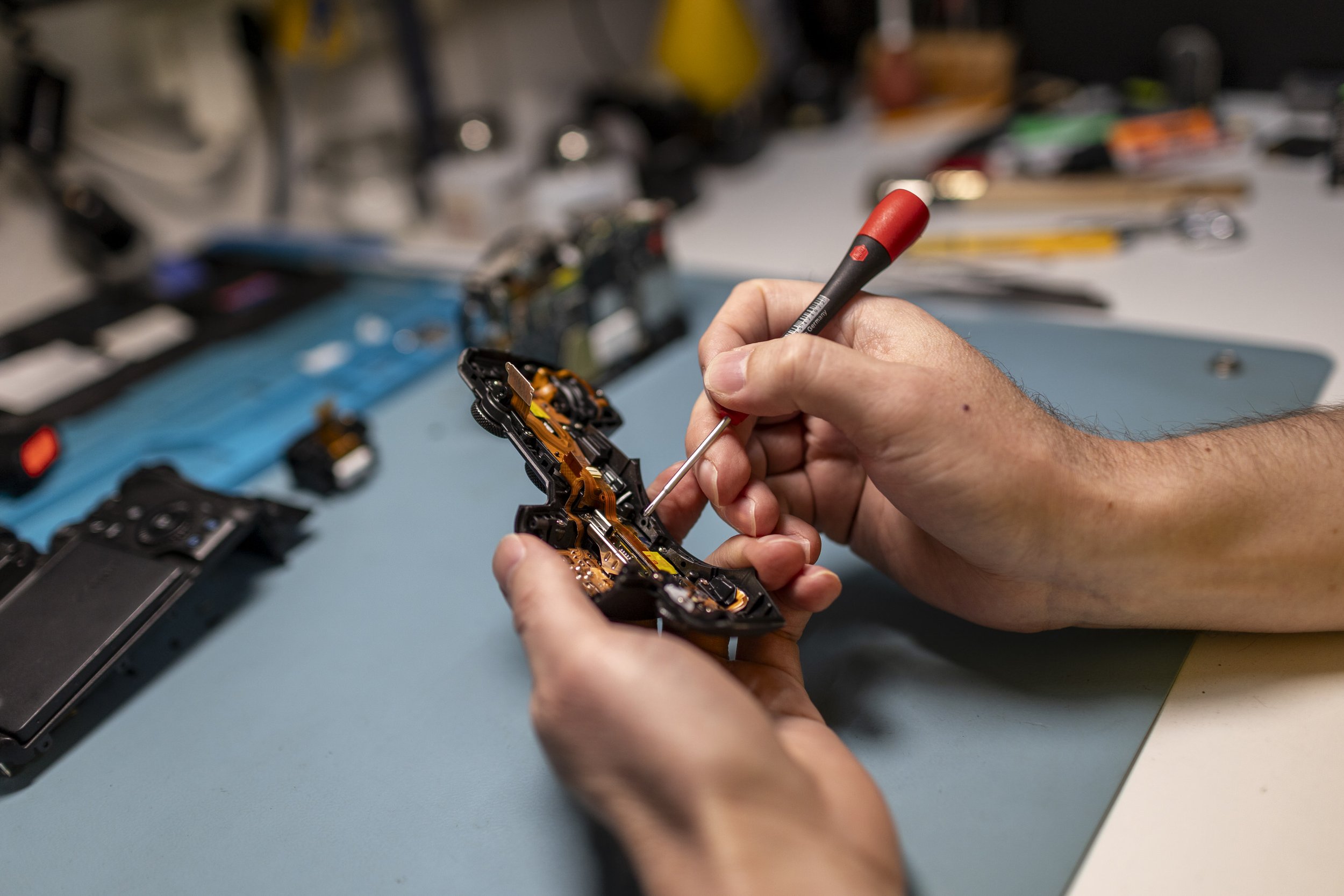 Person repairing an electronic device with a small screwdriver, with various tools and electronic components on a workbench in the background.