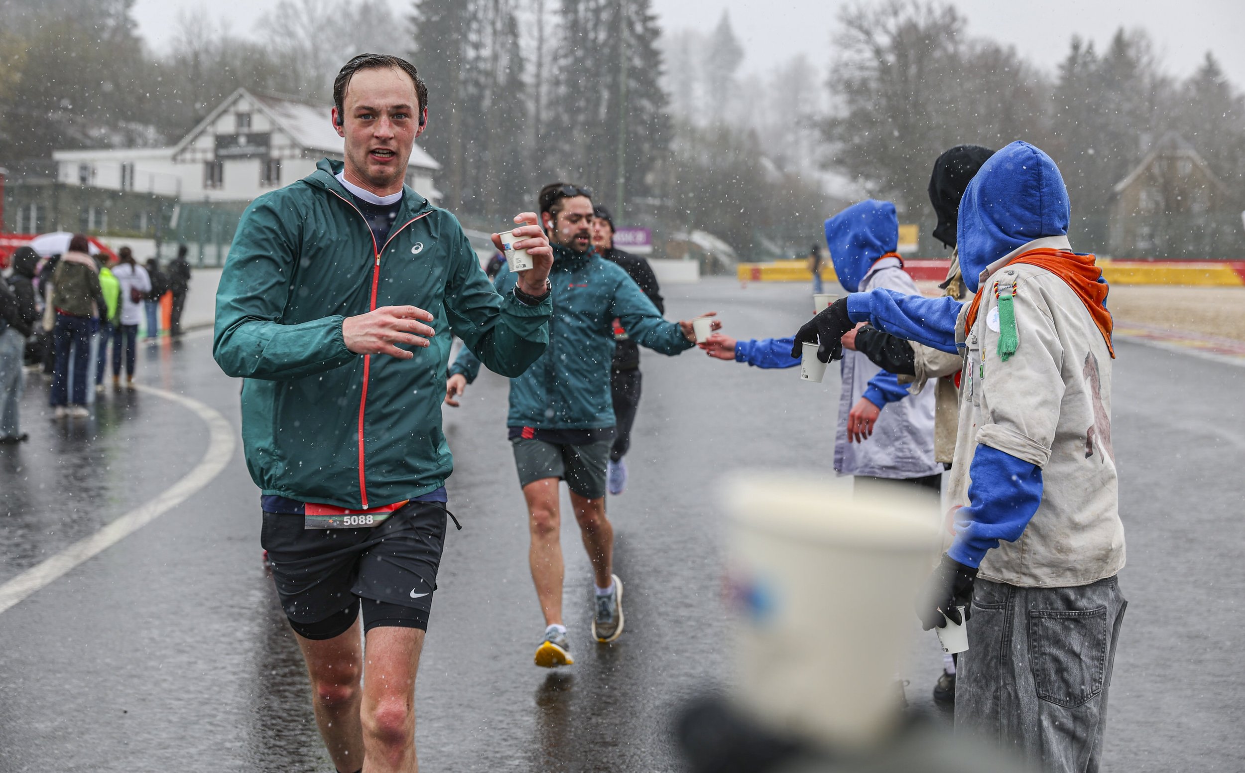 Marathon runner in black shorts and teal jacket running on wet, rainy street, while volunteers hand out beverages in a race event with onlookers in the background and trees and houses on a cloudy day.
