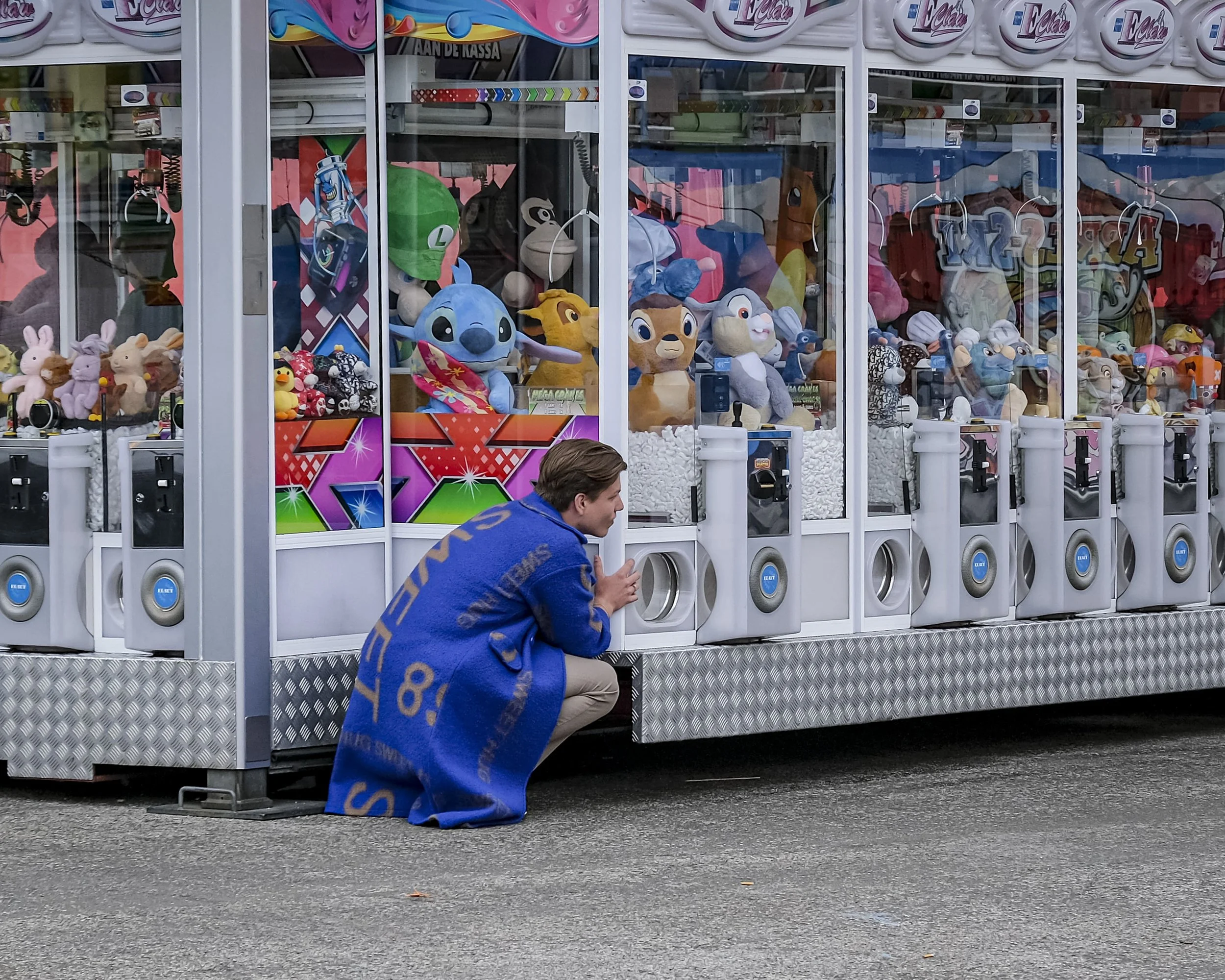 A man crouching in front of a toy claw machine filled with plush toys including characters like Stitch from Lilo & Stitch, Simba from The Lion King, and other stuffed animals. The man is wearing a blue hoodie with orange and yellow lettering on the b