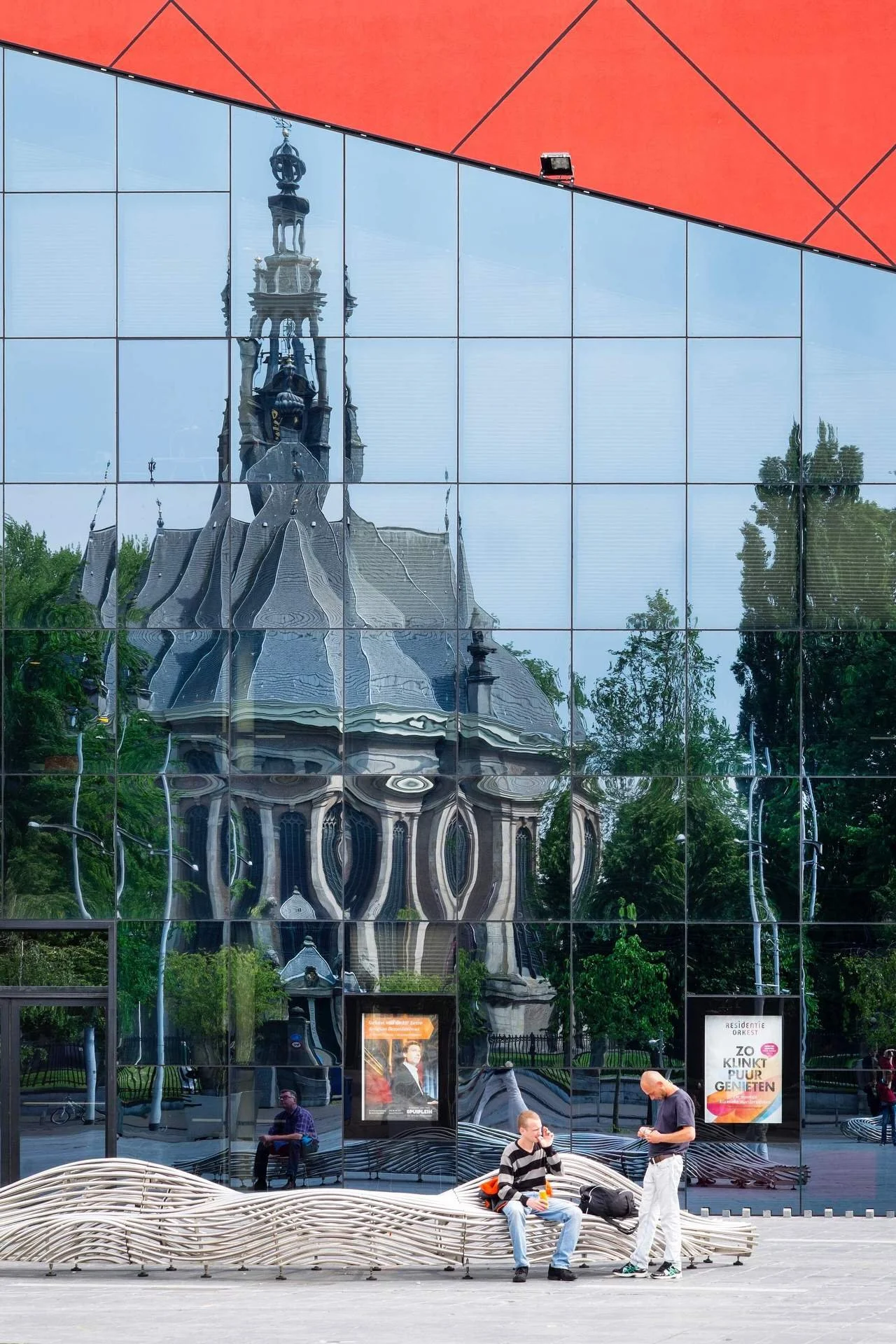 Reflection of an ornate church spire with a clock on a building with a glass facade in an urban park setting, with people sitting on a wavy bench in front.