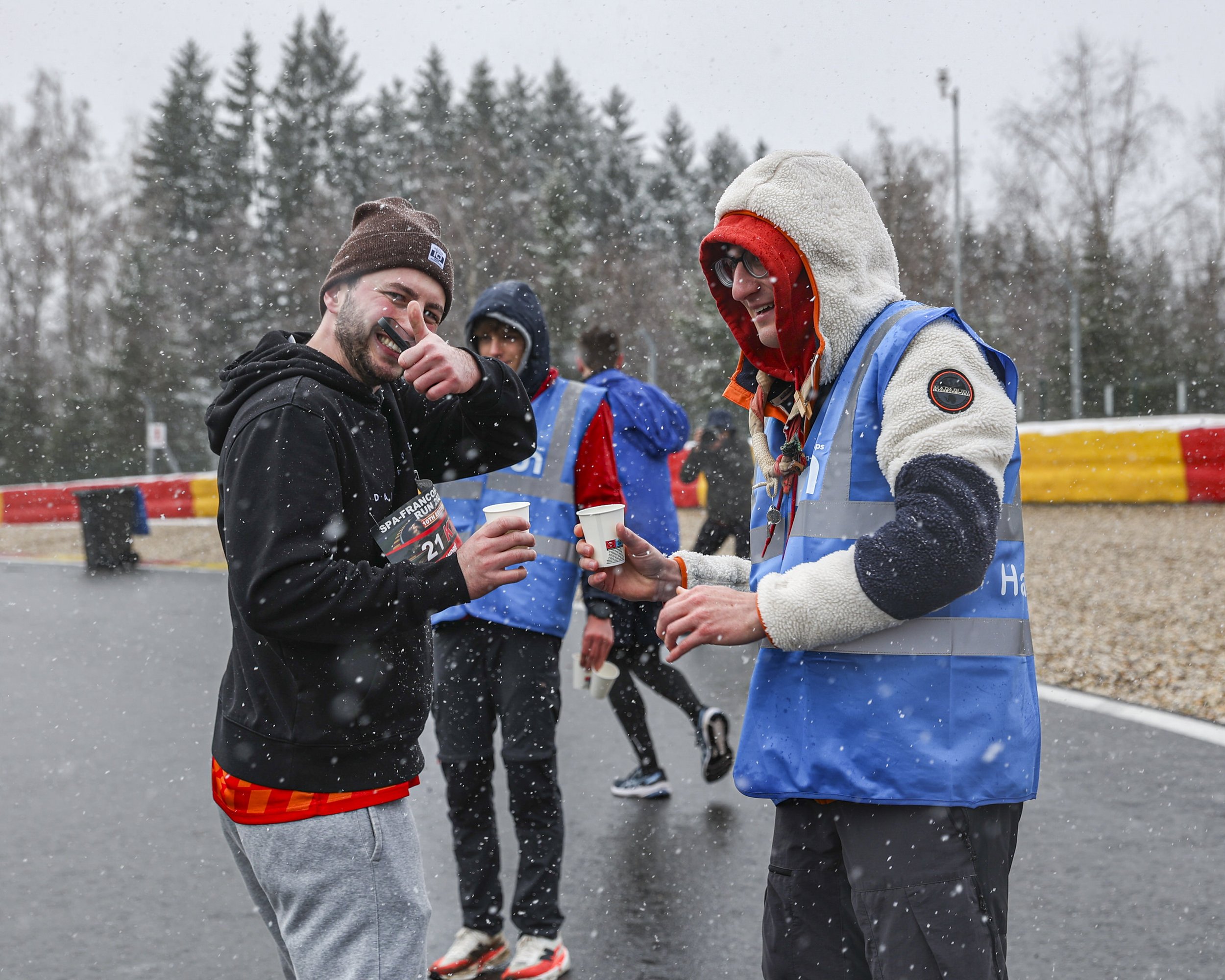 Two men shaking hands at an outdoor event in the snow, with other people in the background. One man is wearing a black hoodie and red shorts, while the other is dressed in a fleece jacket with a hood and glasses. They are smiling and holding cups.