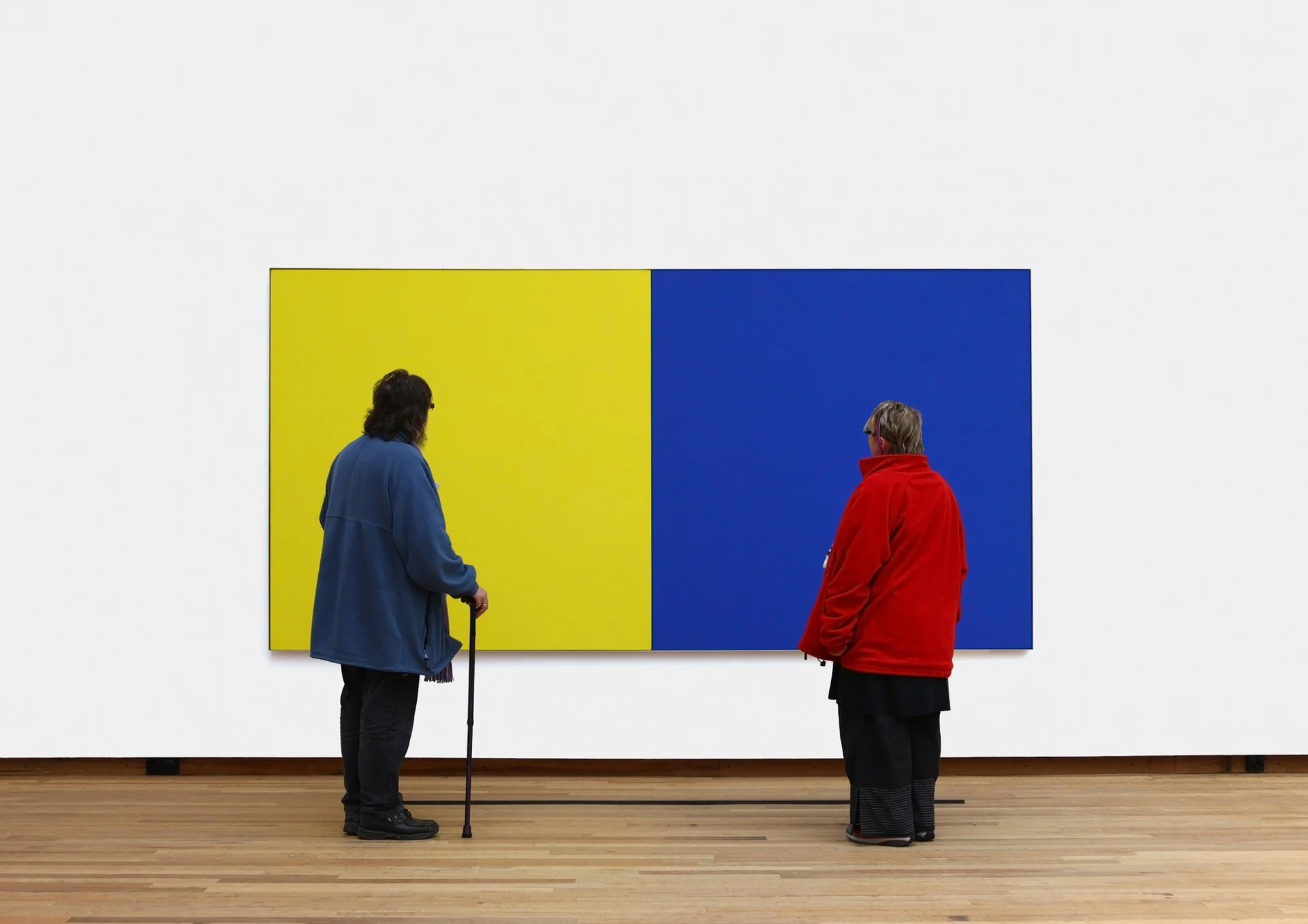 Two elderly women are standing in front of a large artwork with two colored panels, one yellow and one blue, in an art gallery. One woman is using a cane and wearing a blue coat, while the other woman is dressed in a red coat.