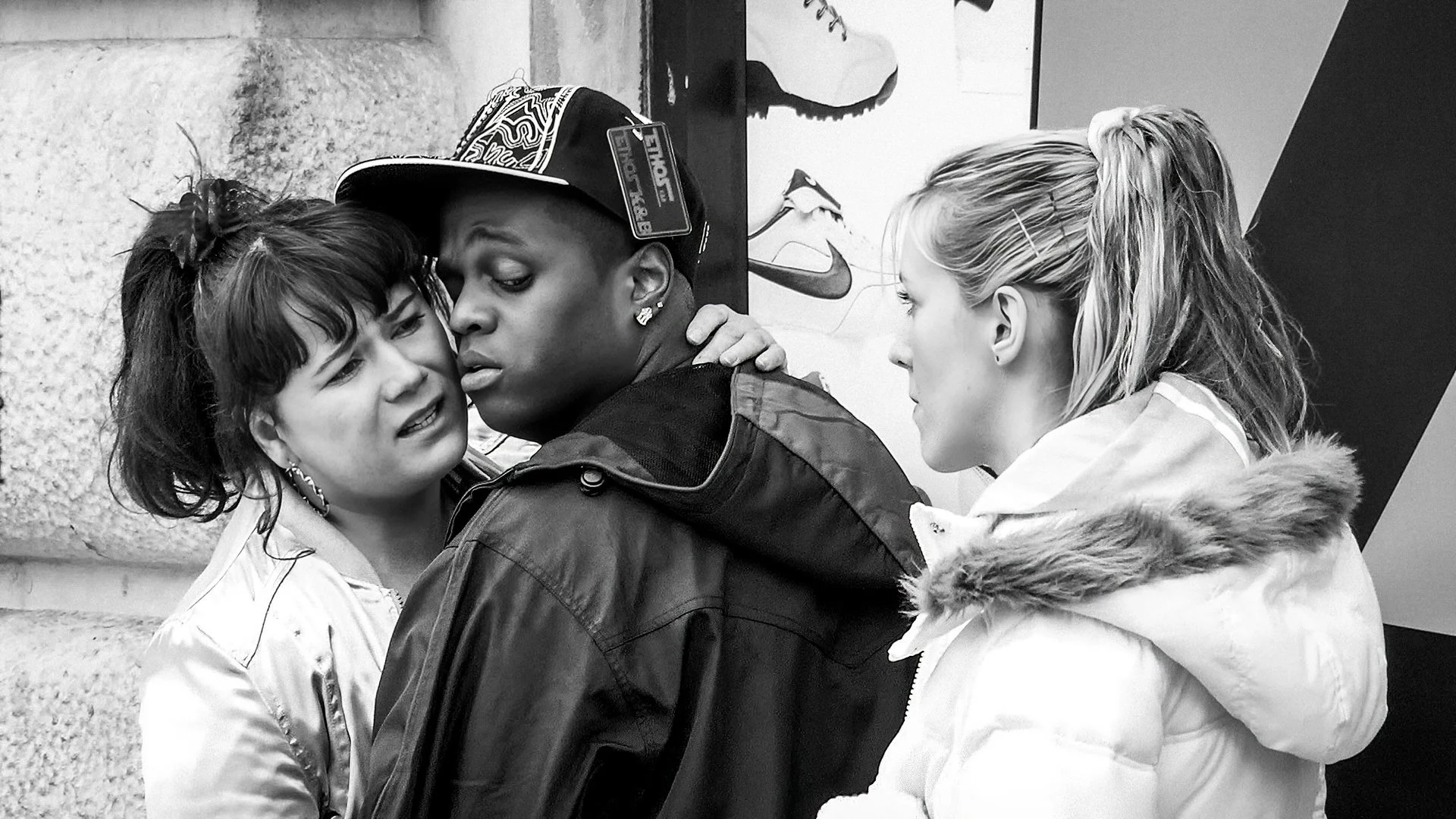 Three women and one man are engaged in a close, serious conversation outside. The women are facing each other, while the man, wearing a cap, is in the middle. The scene is in black and white.