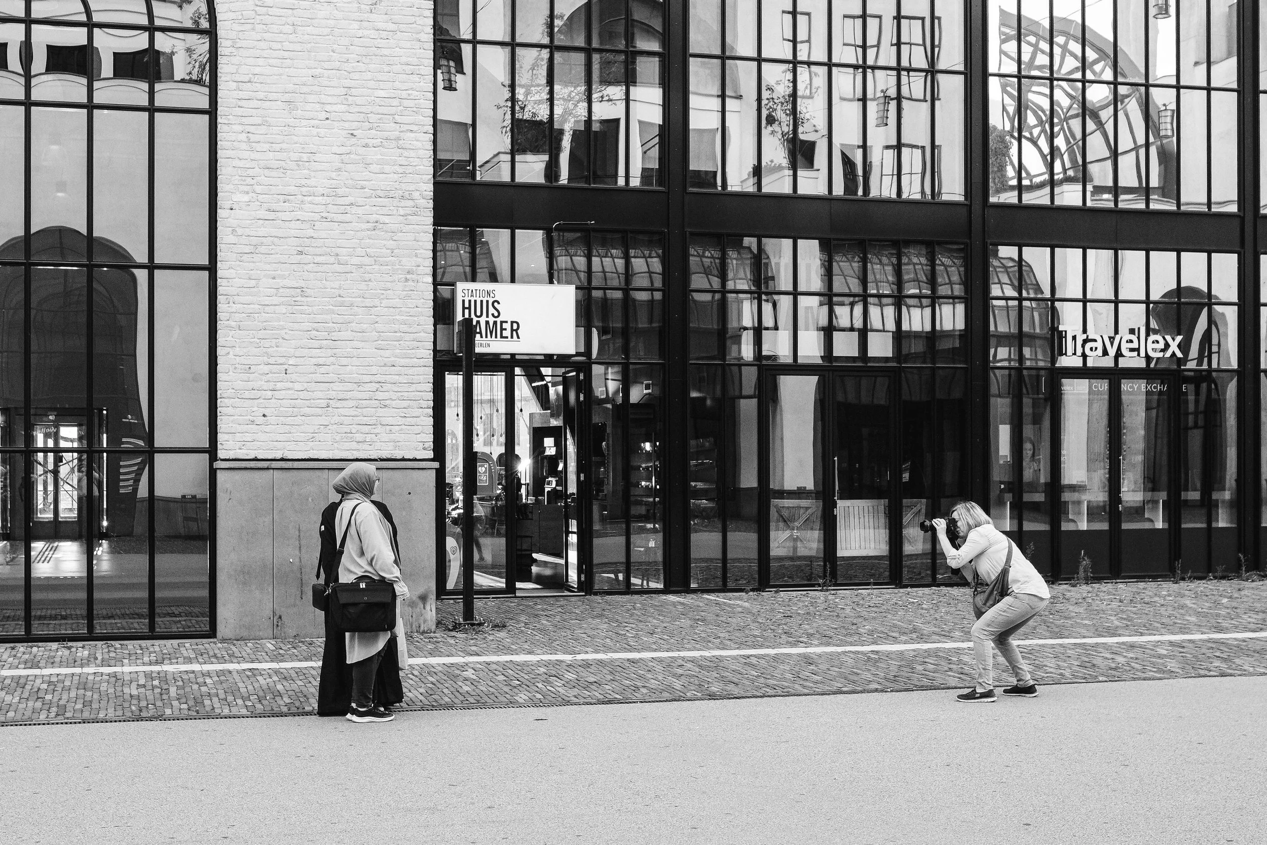 Black and white photo of a modern glass building with reflections, with two women in front. One is standing with a suitcase, the other is crouching taking a photo.