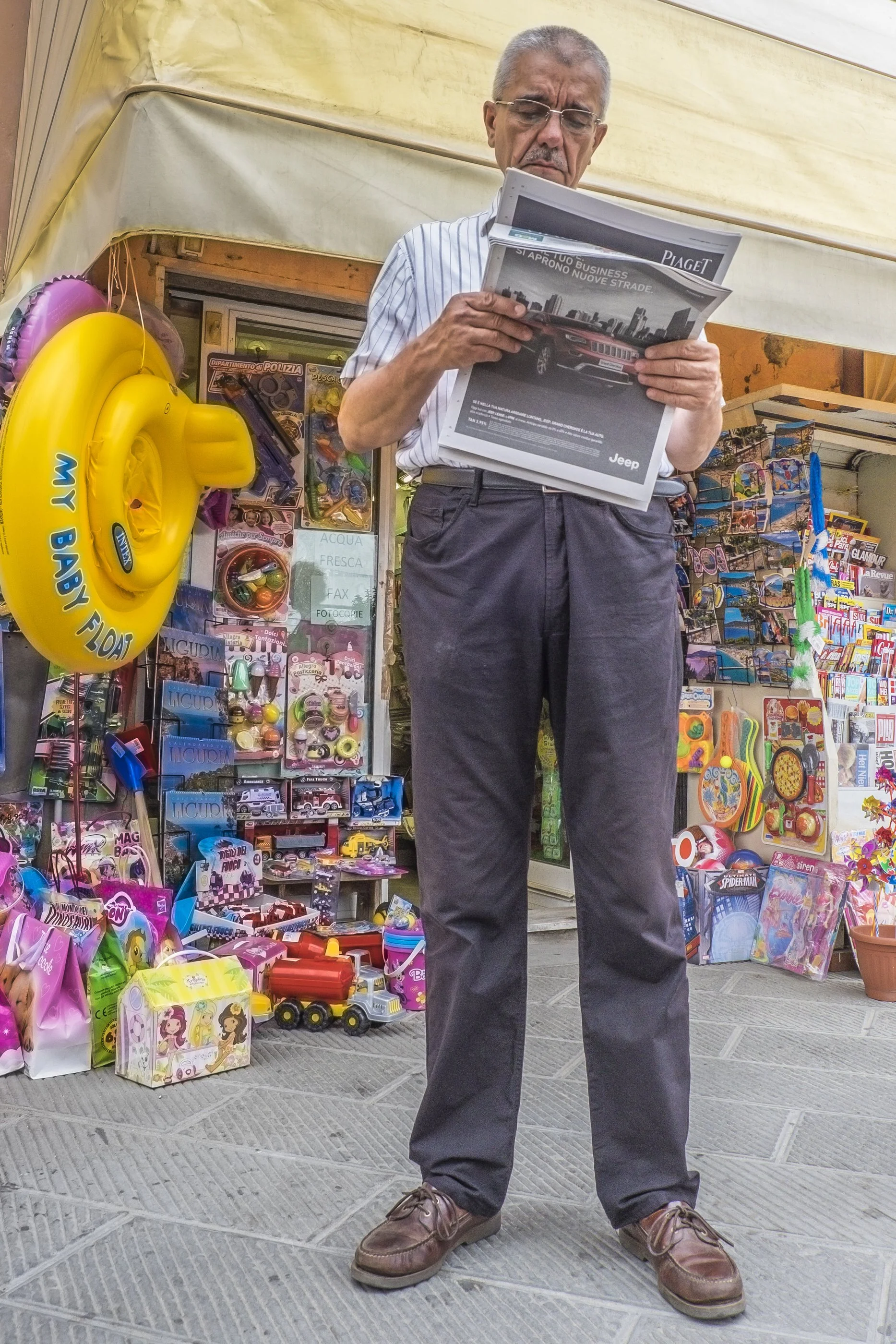 A man in a striped shirt and dark pants stands on a sidewalk reading a newspaper. Behind him is a street vendor stall with colorful toys and accessories, including a yellow inflatable swim ring labeled 'MY BABY FLOAT.'