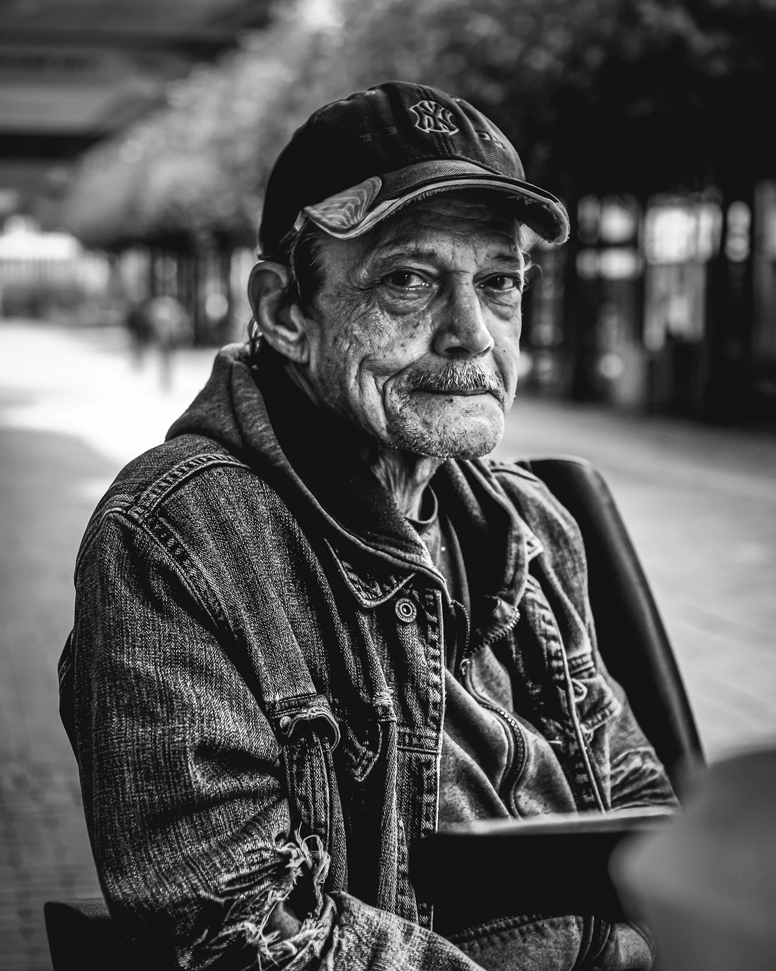 A black and white photo of an elderly man with a cap, sitting outdoors at a park or street, looking back at the camera.