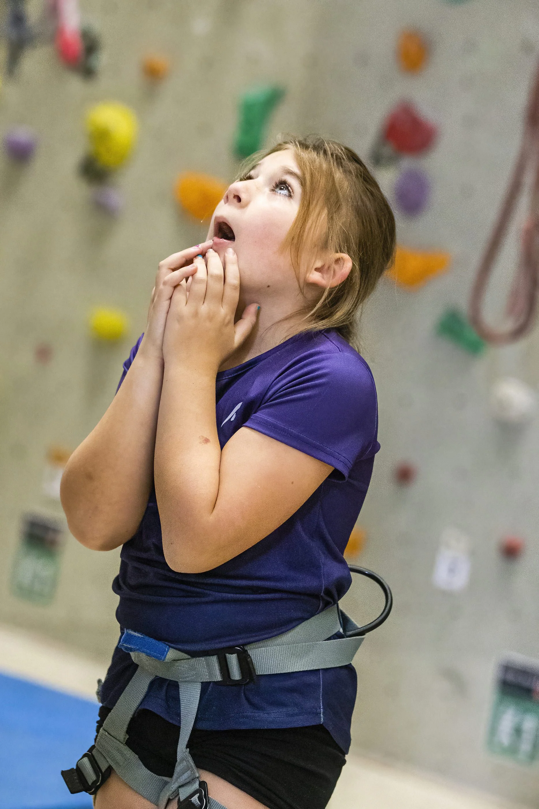 A young girl in climbing gear at an indoor rock climbing gym, wearing a harness and looking up at the climbing wall with colorful holds.