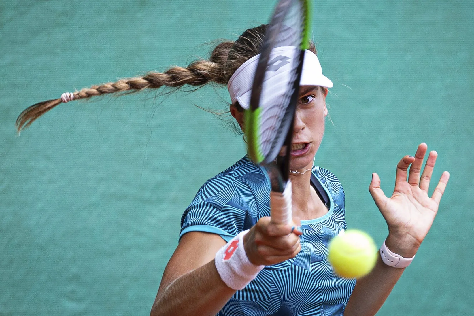 A female tennis player with braided hair, wearing a white visor and blue sports shirt, is hitting a tennis ball with her racket during a match. Her expression is intense and focused, with a green textured background.