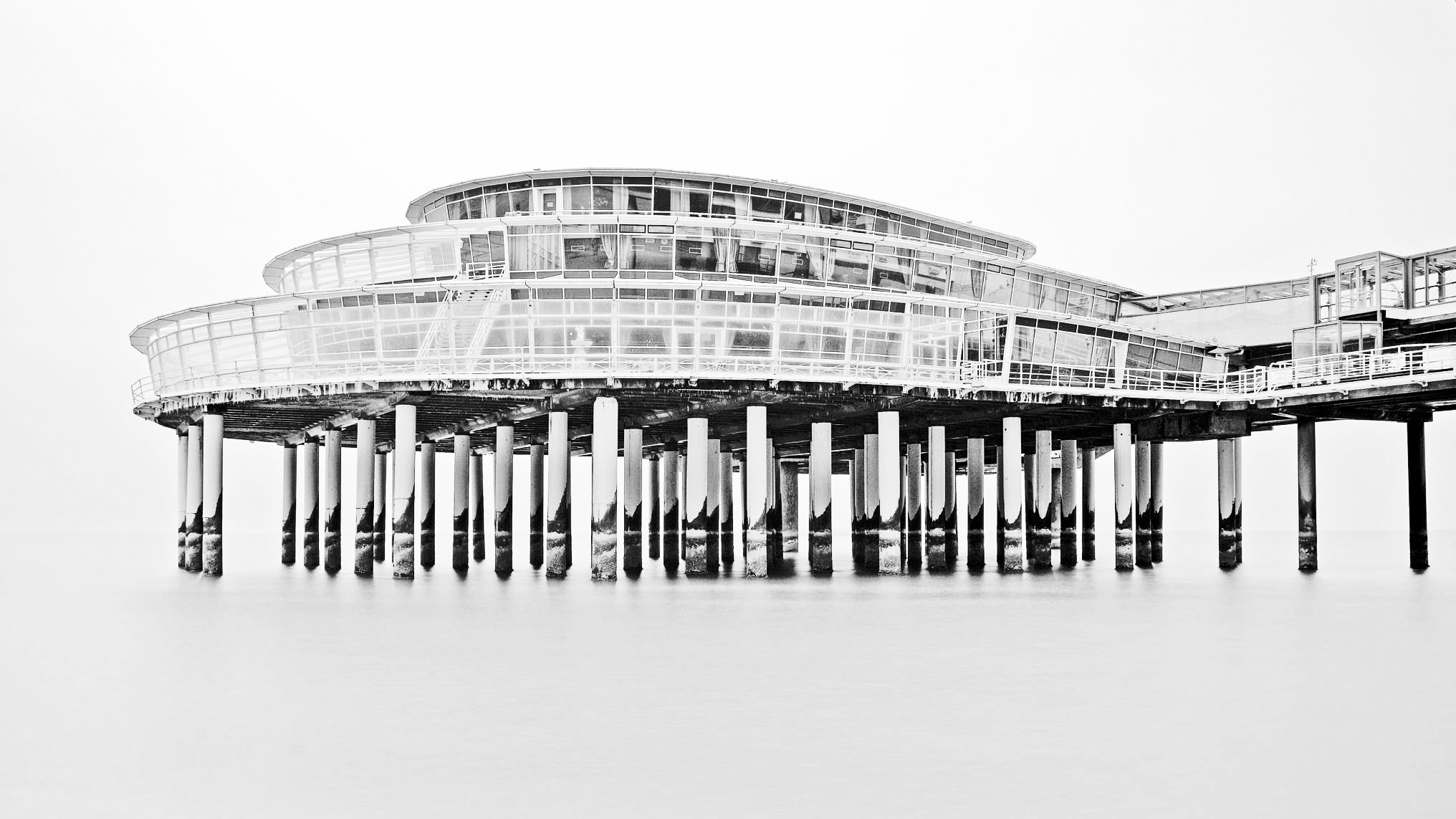 A black and white photograph of a modern, multi-level building on stilts over water, with large glass windows and curved architectural design.