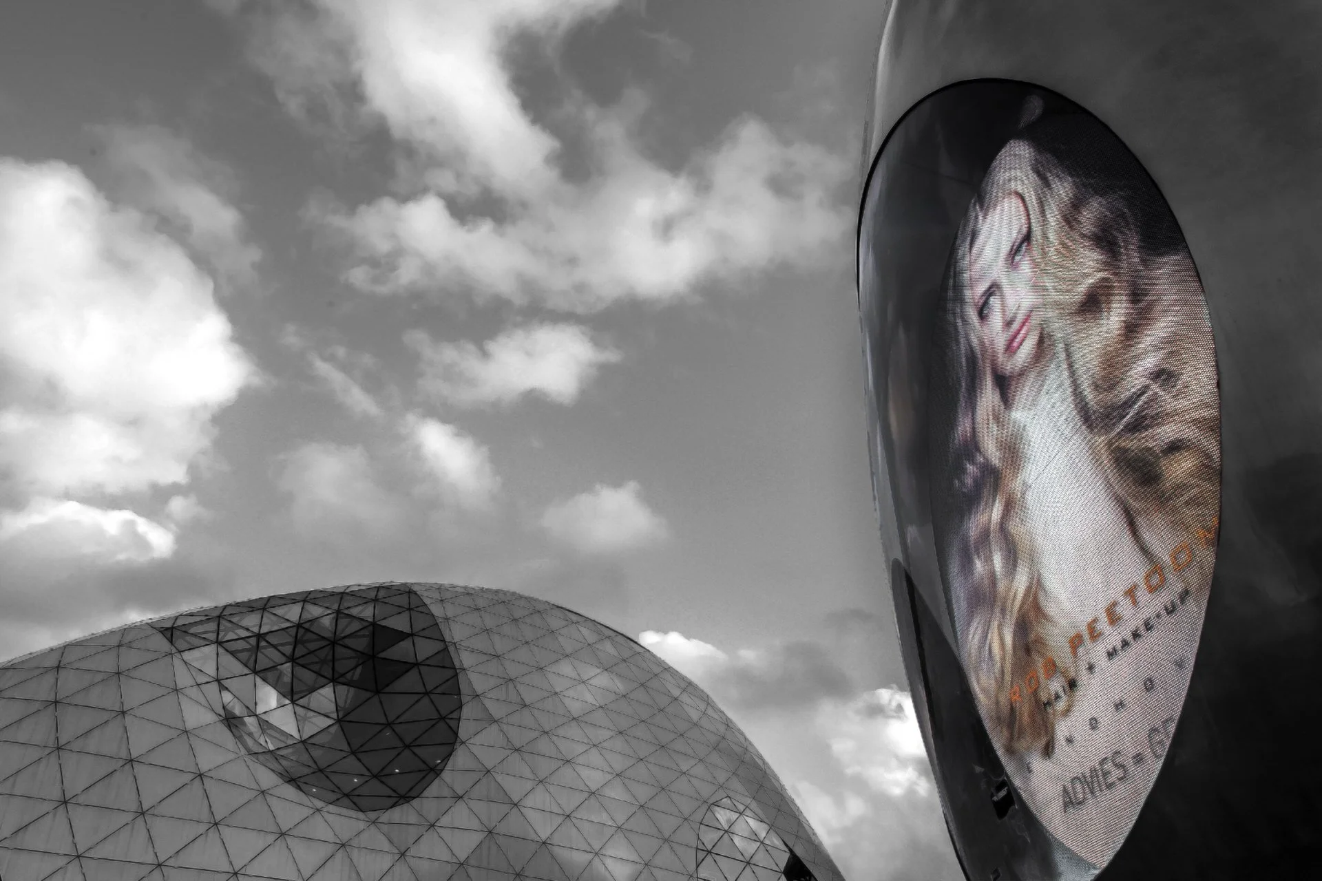 A modern sports stadium with a geodesic dome design and a large digital billboard displaying a person's face against a cloudy sky.