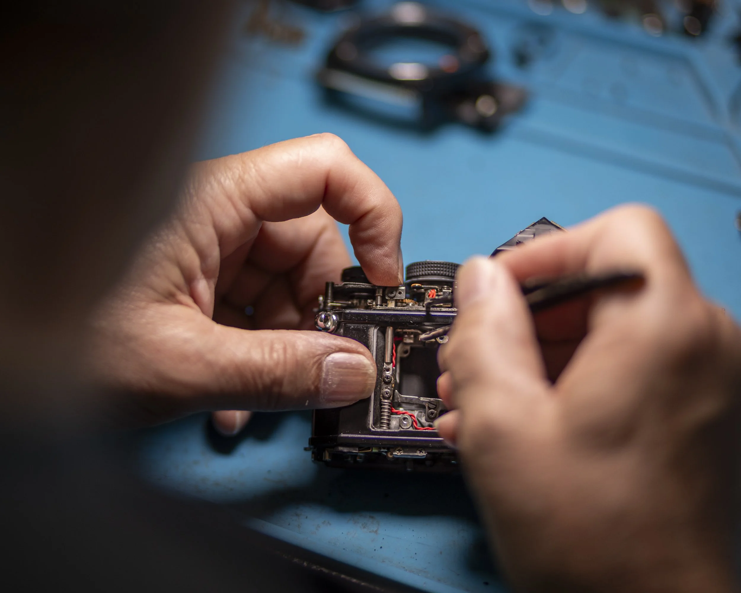 Close-up of hands working on an electronic device, possibly a small robot or circuit board, on a blue surface with blurred tools or parts in the background.
