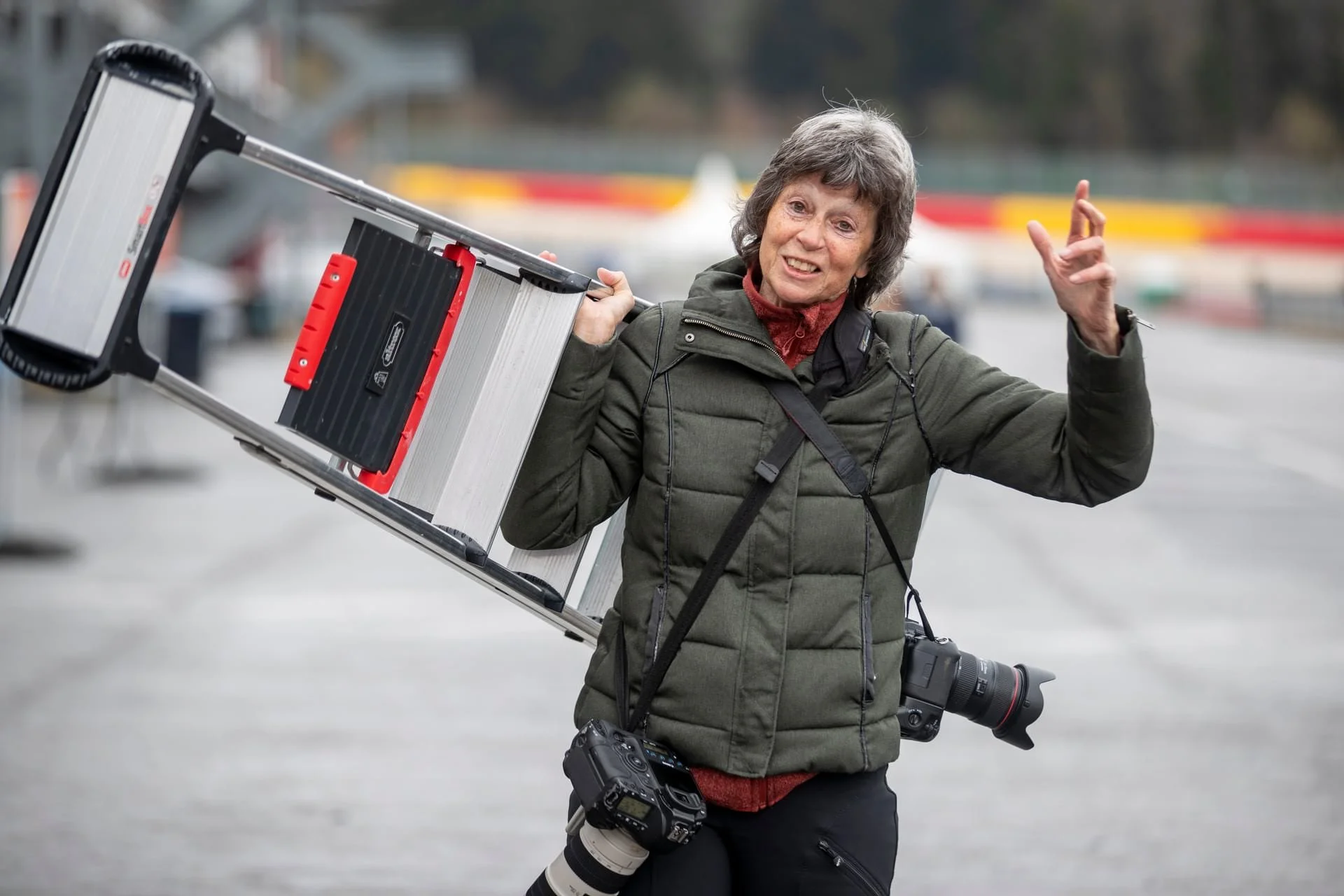 A woman carrying photography equipment, including a ladder with a projector mounted on it, camera lenses, and a camera, outdoors with blurred background.