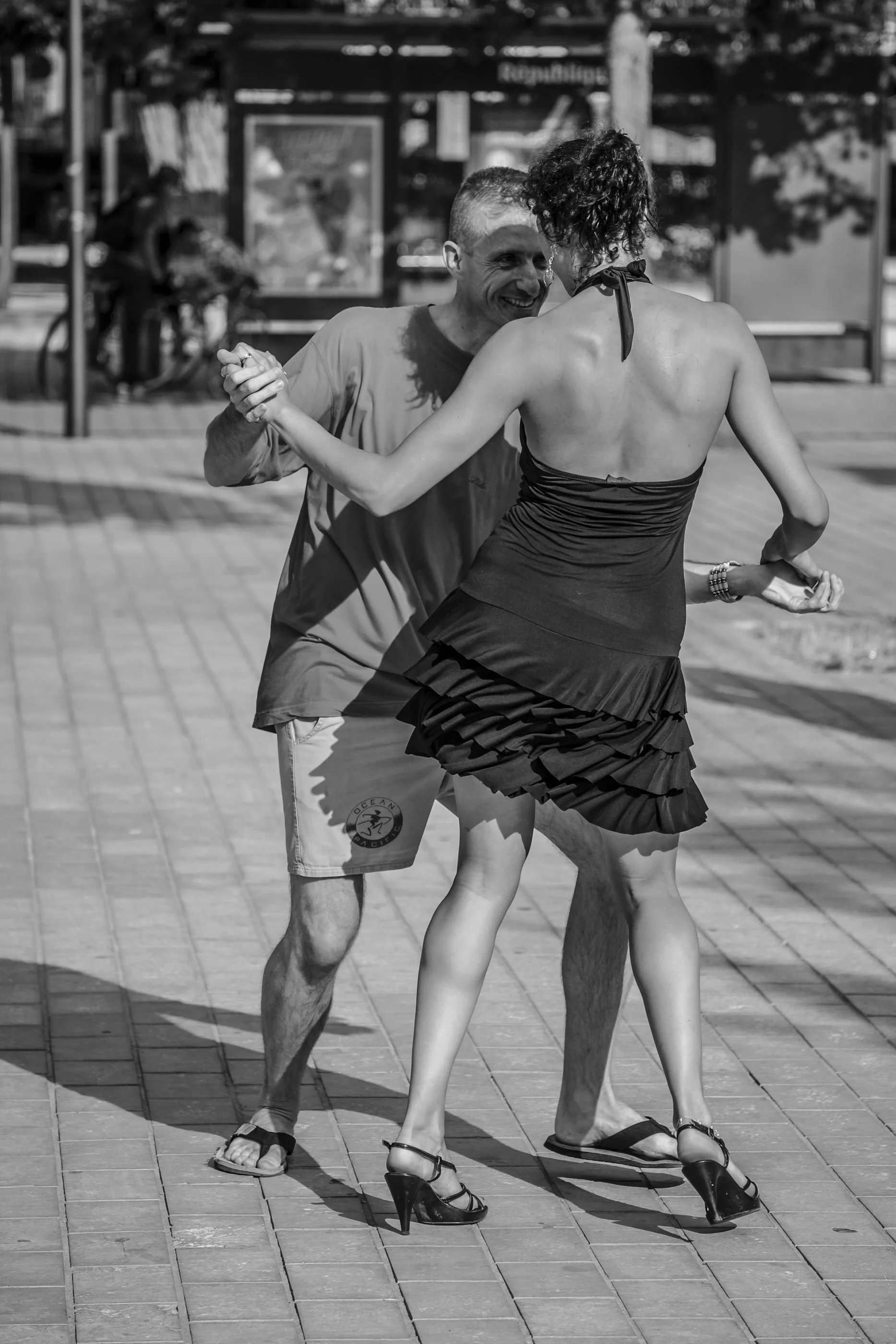 A man and woman dancing closely outdoors, with the man smiling and the woman in a black dress wearing high heels, visible in a street scene with a bike and a public noticeboard in the background.