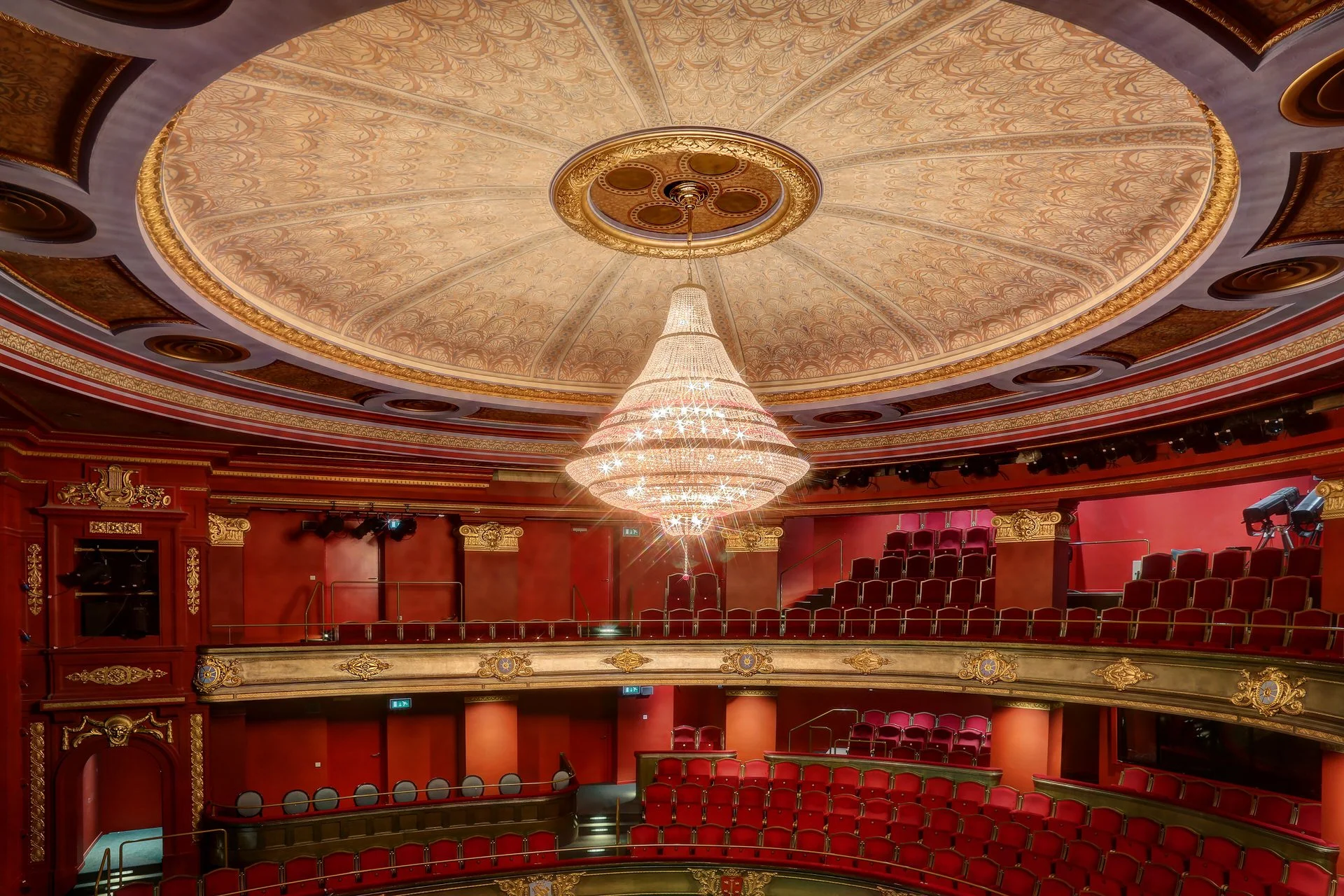 The interior of a grand theater with a domed, ornately decorated ceiling featuring gold accents and a chandelier hanging in the center. The theater has red velvet seats and elaborate red and gold decor.
