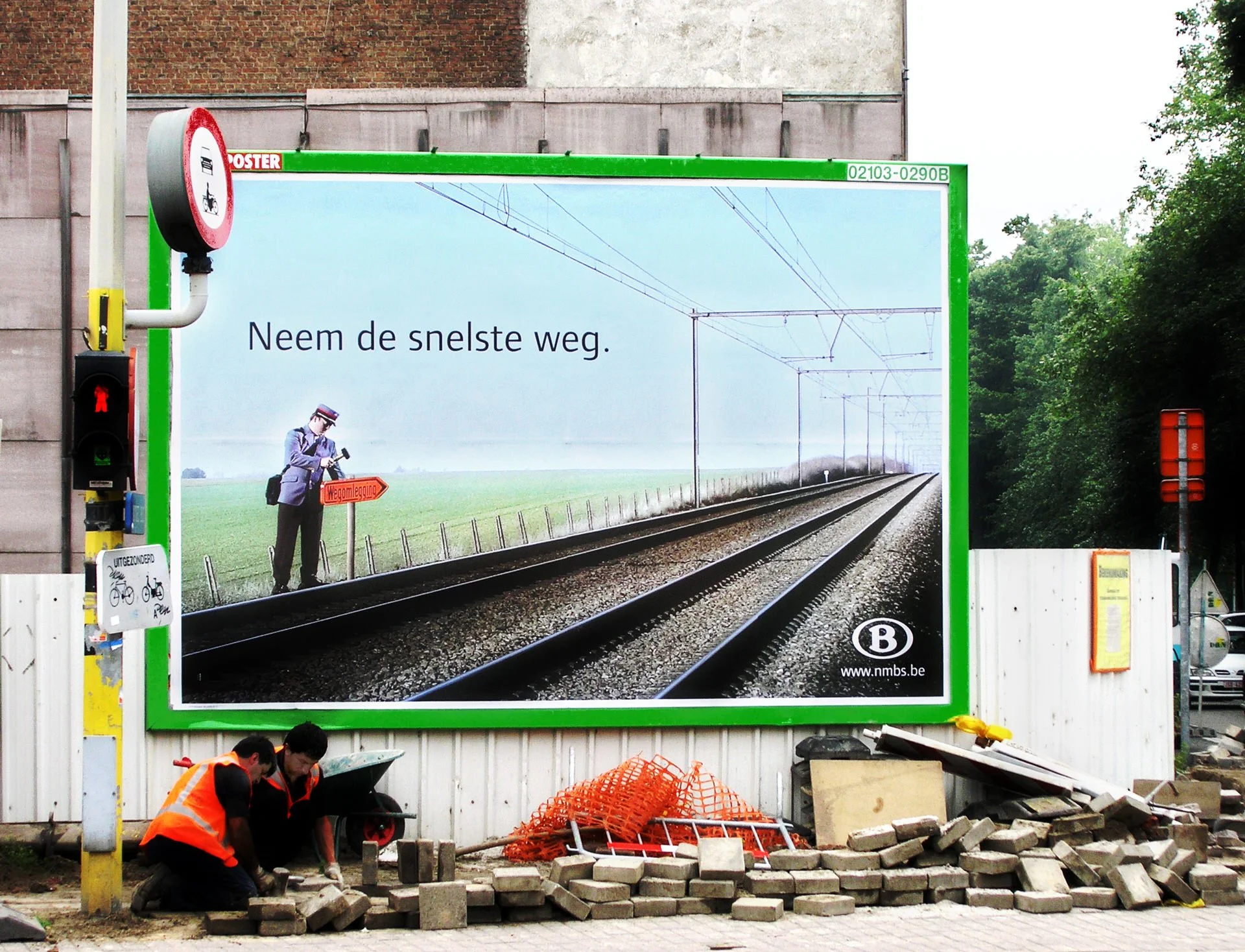 Billboard with Dutch text and train tracks, two construction workers laying bricks in front, and construction tools and materials nearby.