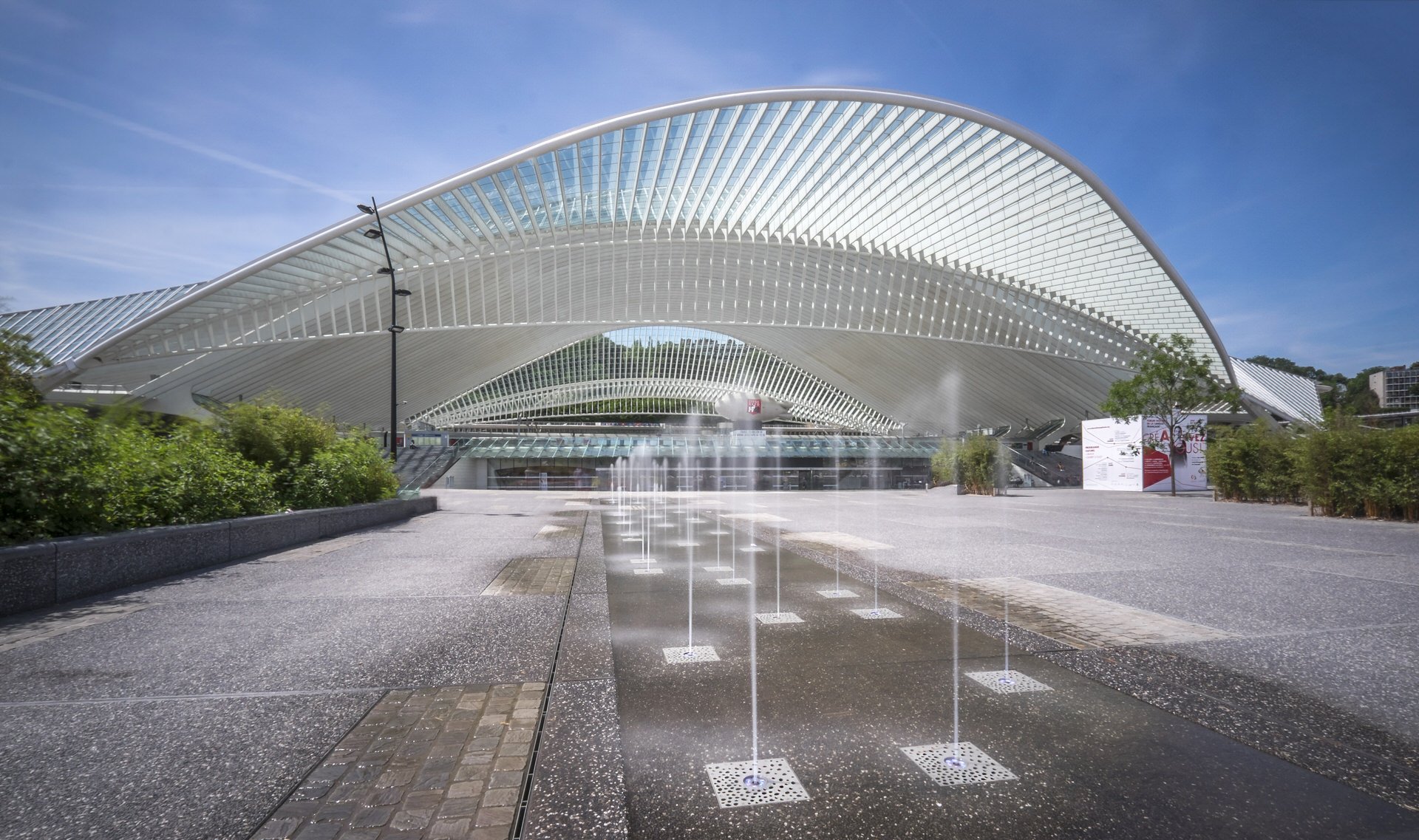 Modern architectural building with sweeping white roof structures, glass facades, and a water fountain feature in the foreground, situated in a landscaped urban area.