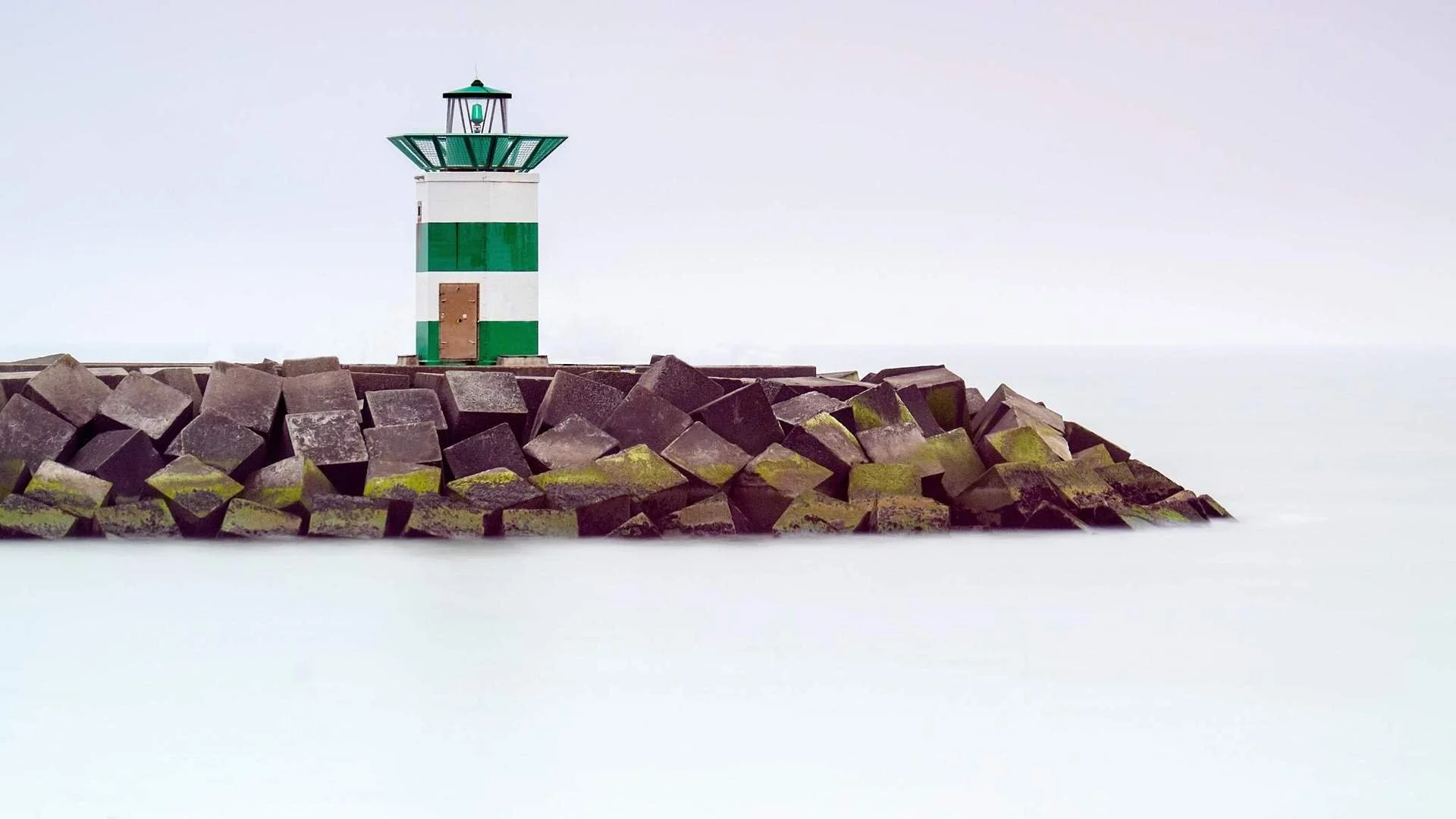 A lighthouse with green and white stripes standing on a rocky breakwater, with the sea and a cloudy sky in the background.