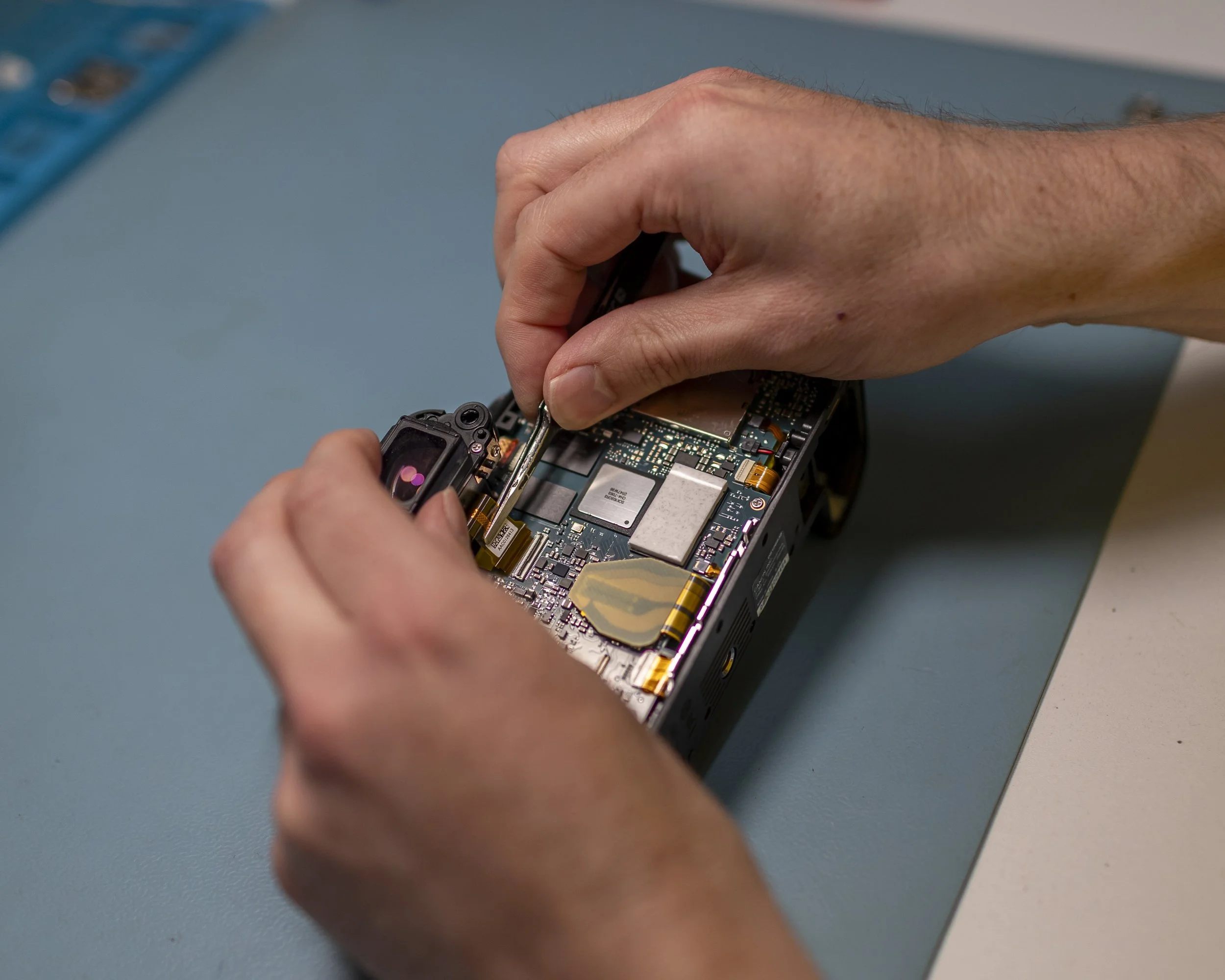 A person's hand working on the internal components of an electronic device, possibly a smartphone or small circuit board, with a blue background surface.