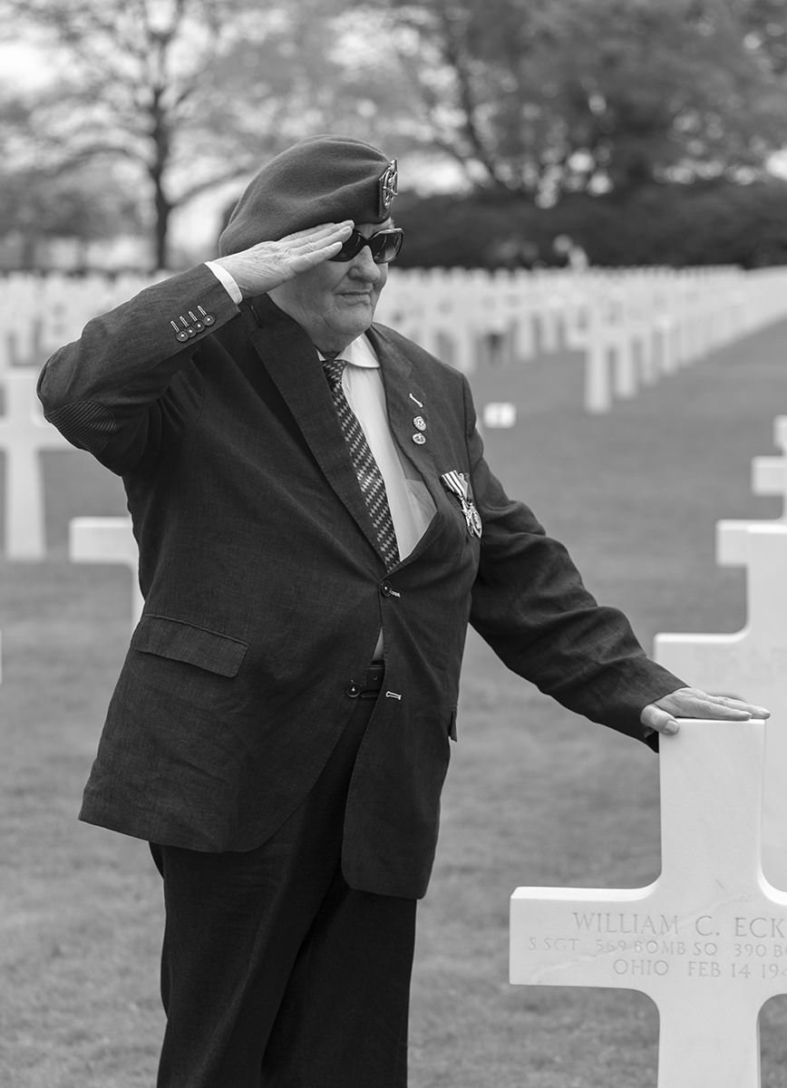 An elderly man in a military uniform saluting at a memorial cemetery, wearing a beret, sunglasses, and several medals, with white crosses in the background.
