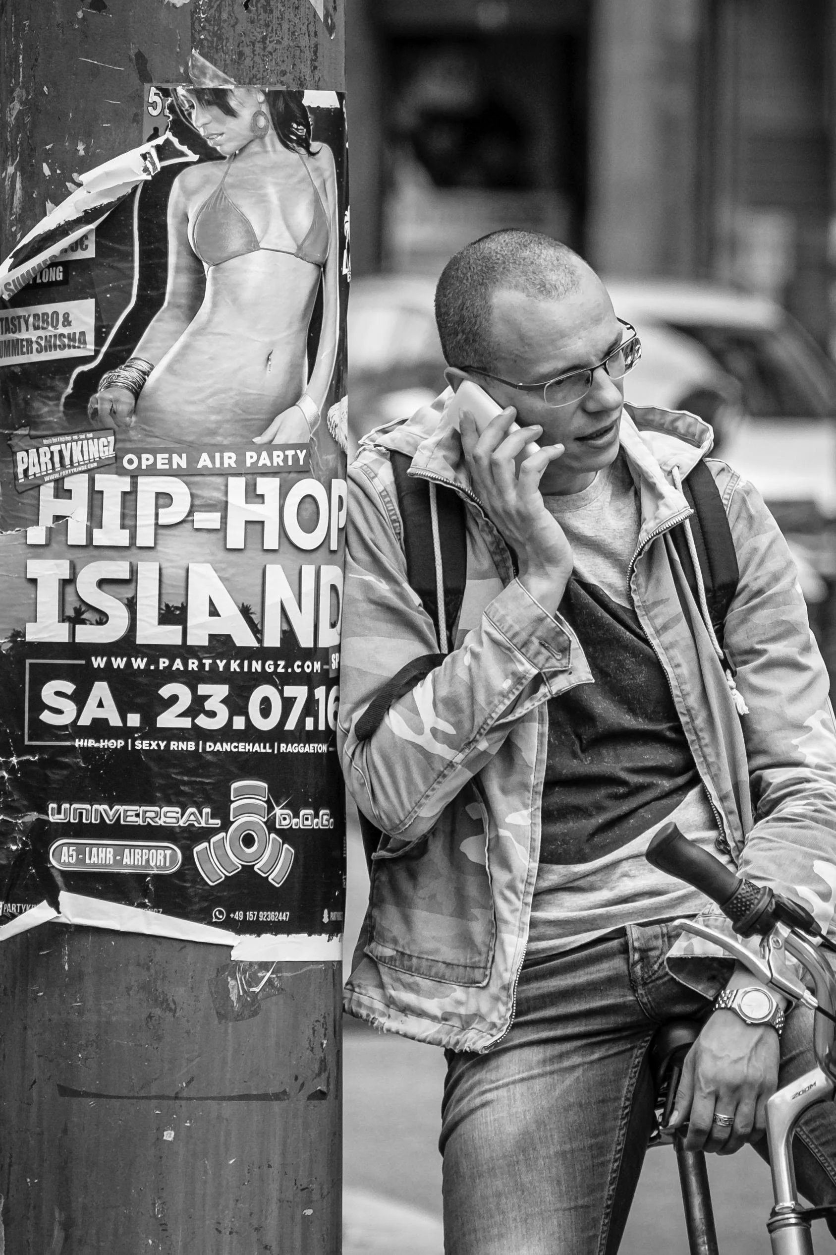 A man in glasses, a striped jacket, and casual clothes is riding a bicycle and talking on a cellphone. There is a poster on a pole next to him advertising a hip-hop event on July 23, 2016, featuring a woman in a bikini on the poster.