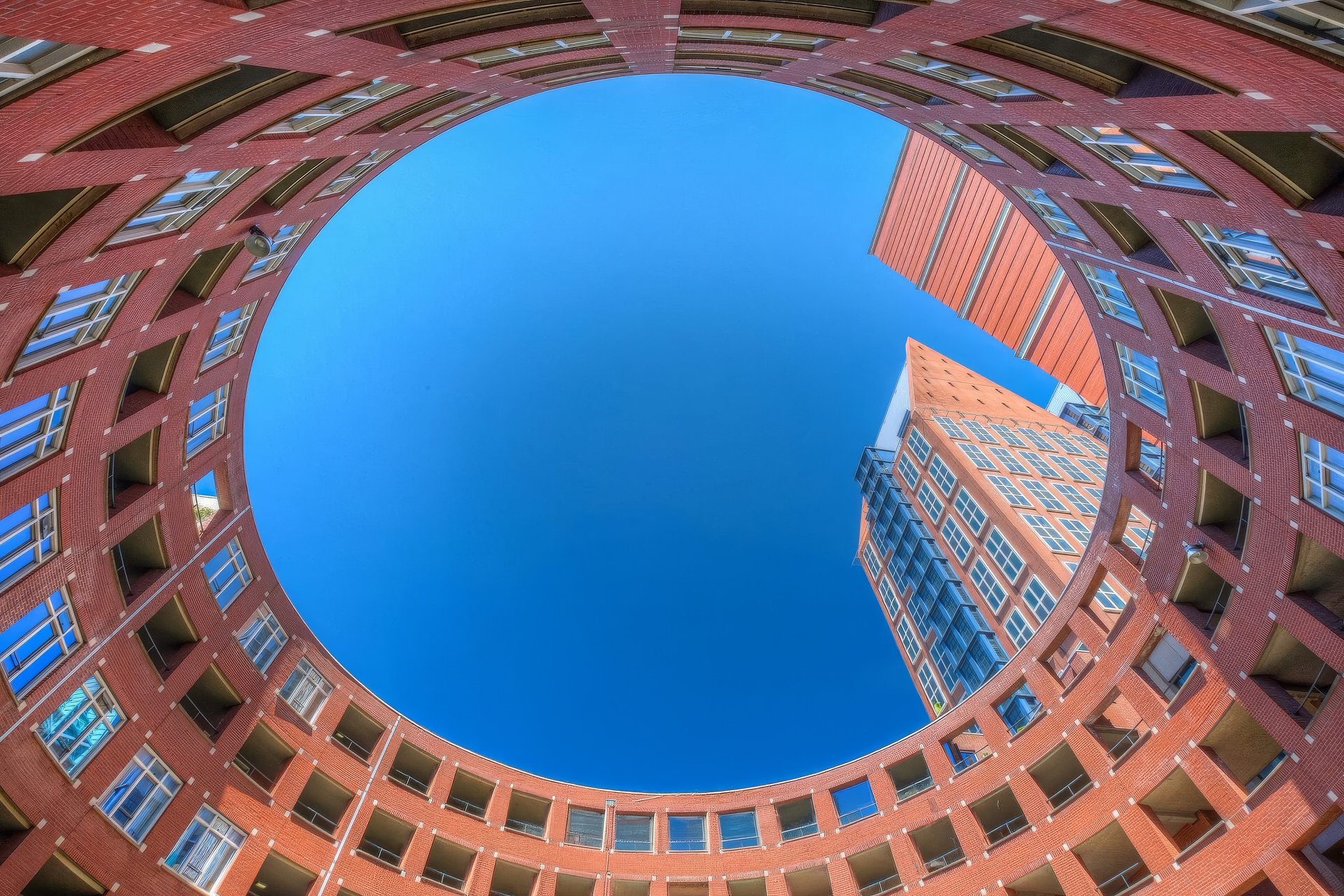 Looking up at a circular apartment complex with red brick exterior, multiple windows, and balconies. The sky and a building are visible inside the circular courtyard.