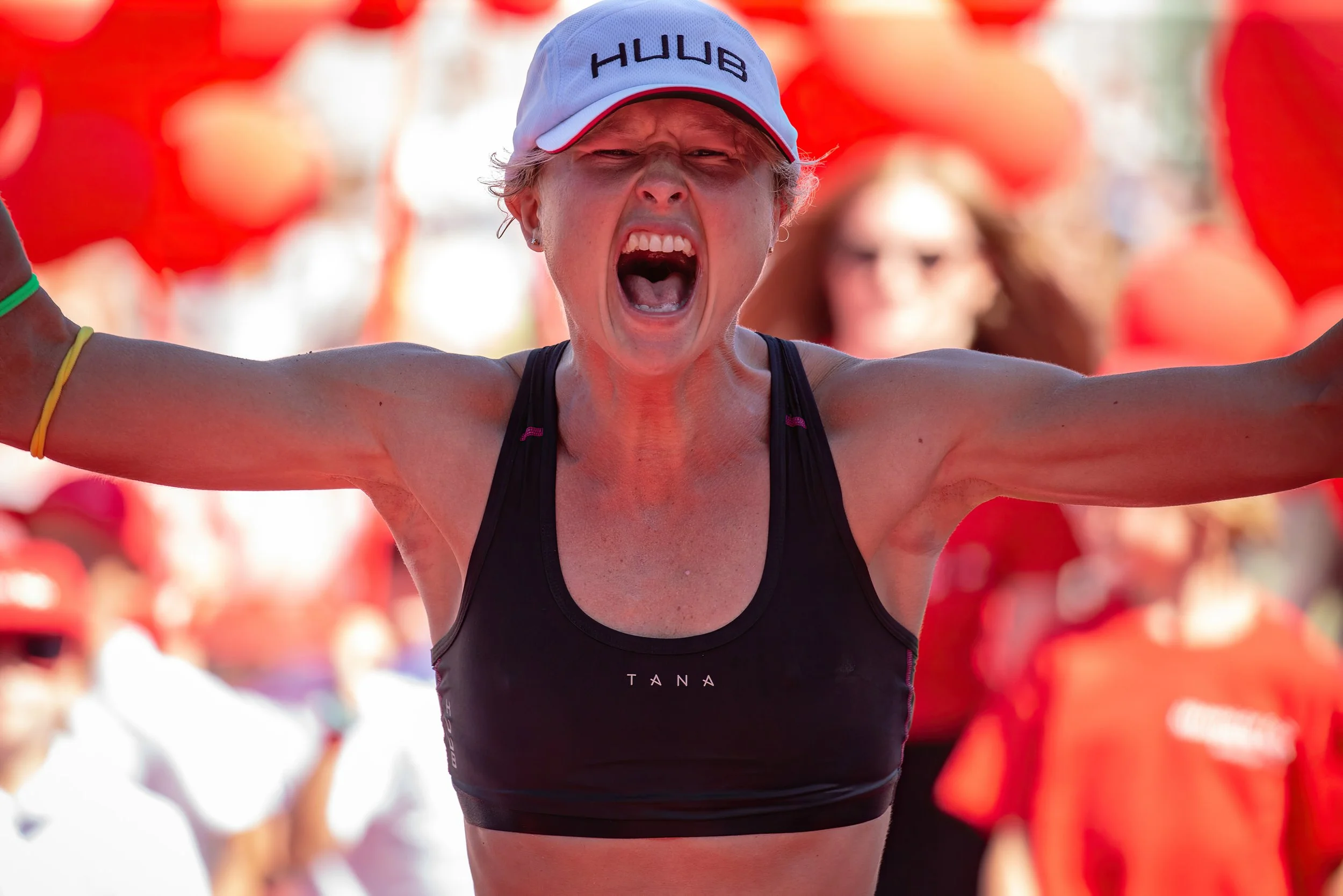 A woman with short blonde hair wearing a white cap with 'HUB' written on it, a black sports tank top with 'TANA' on it, and a yellow wristband, appears to be in a marathon or race with her arms raised, expressing joy or celebration amid balloons.
