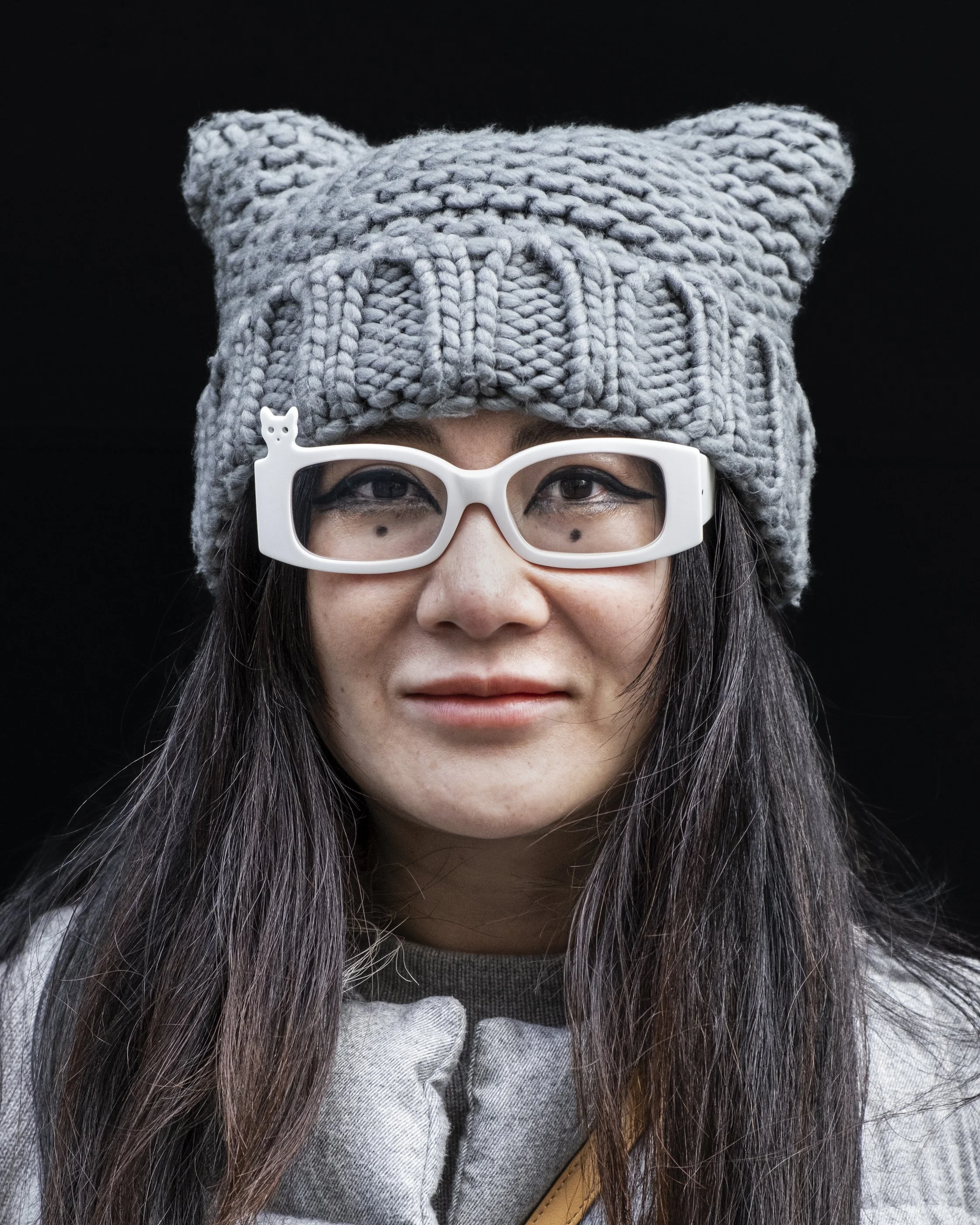 Close-up photo of a person wearing a large, soft, gray knit hat with bear ears, white glasses with a small bear figure on the side, and long dark hair against a black background.
