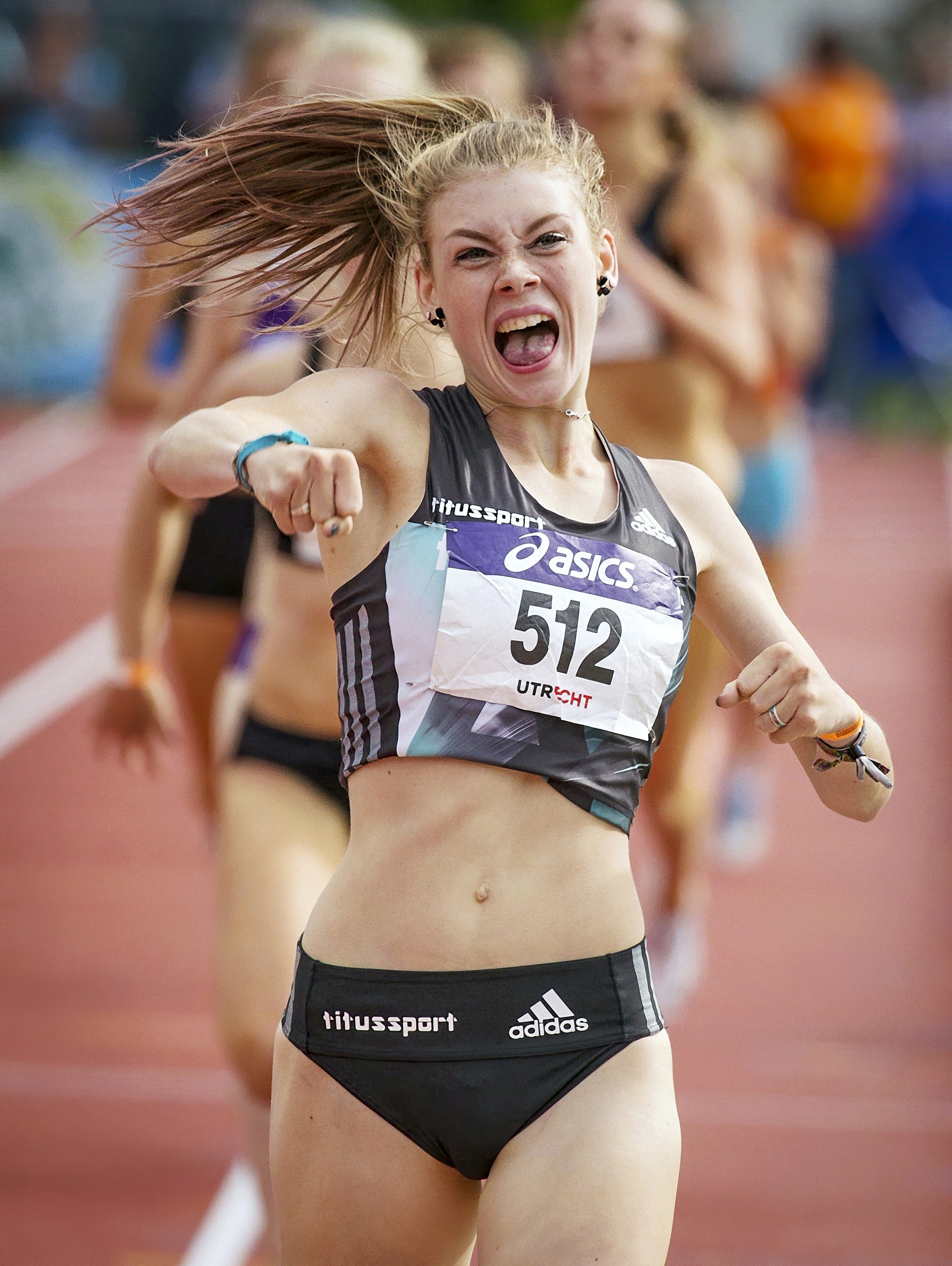 A female athlete running on a track, wearing black athletic shorts and a sports top, with her hair flying and an expression of determination. The background shows other runners and a red track surface.