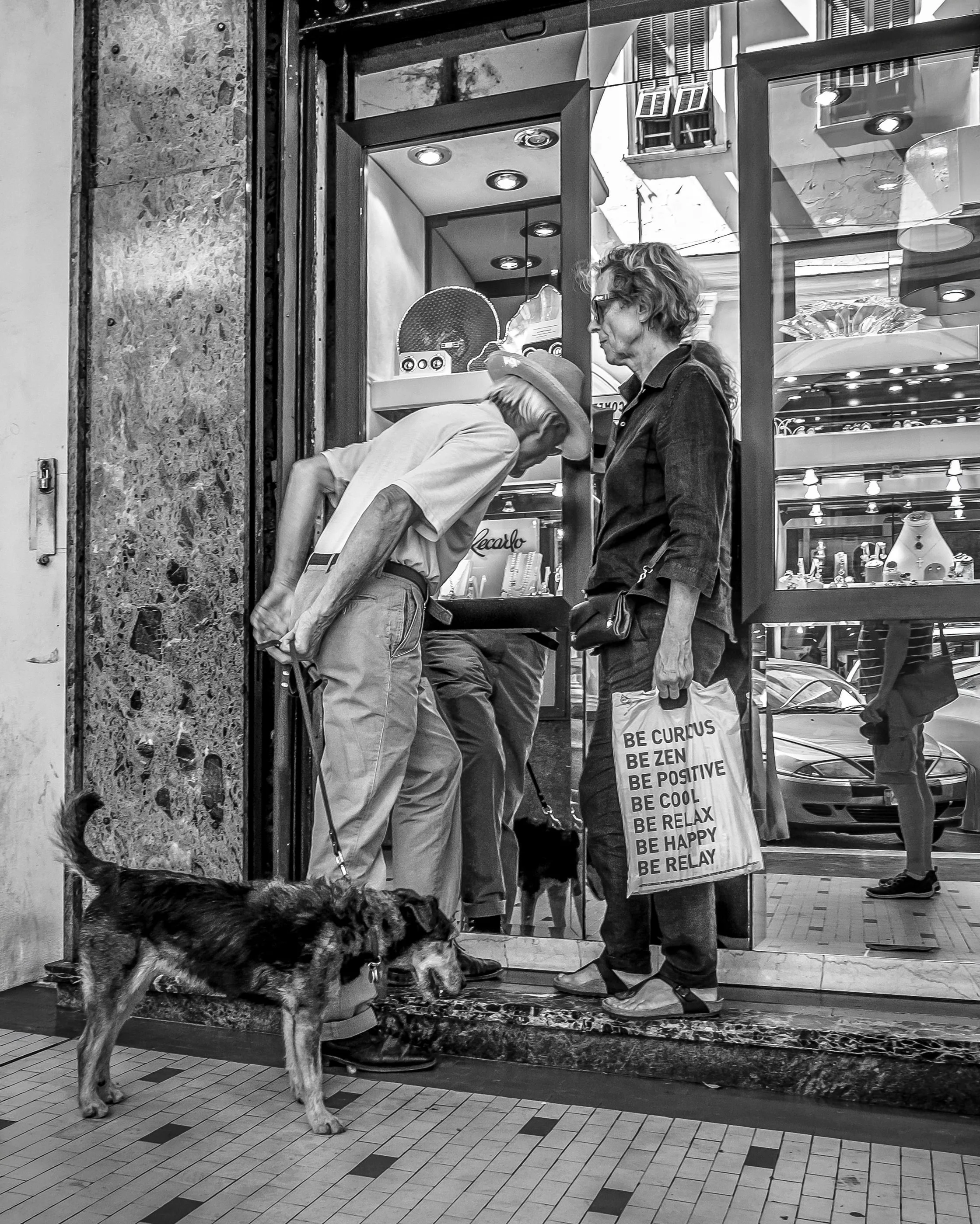 A man in a cap and a woman in glasses and a jacket look at a dog in front of a store window. The woman holds a shopping bag with inspirational messages. The store window displays various items, and the street is visible with reflections in the glass.