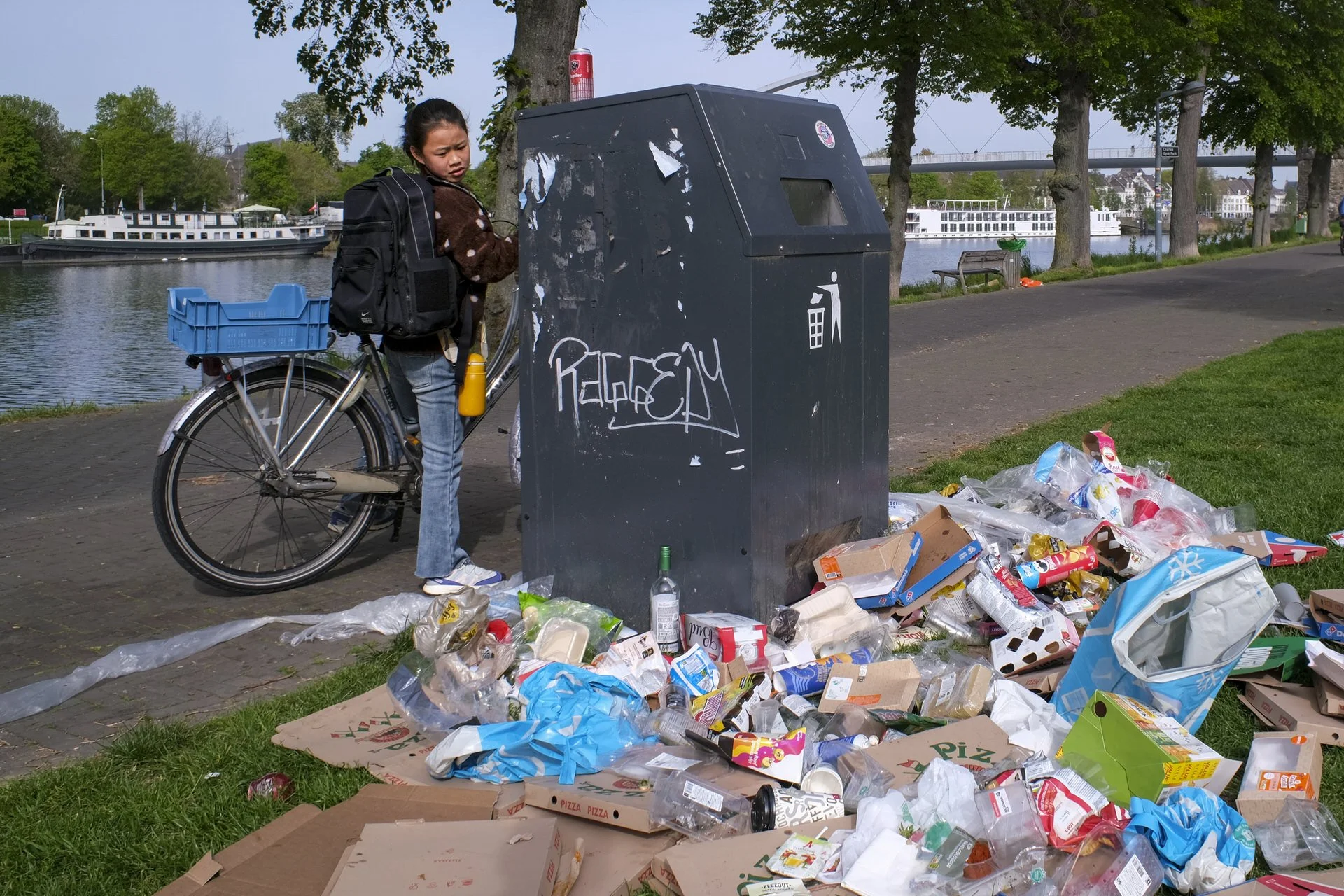A young girl with a backpack stands by a large overflowing trash bin filled with spilled garbage, including pizza boxes, bottles, and plastic packaging, near a river with boats and trees.
