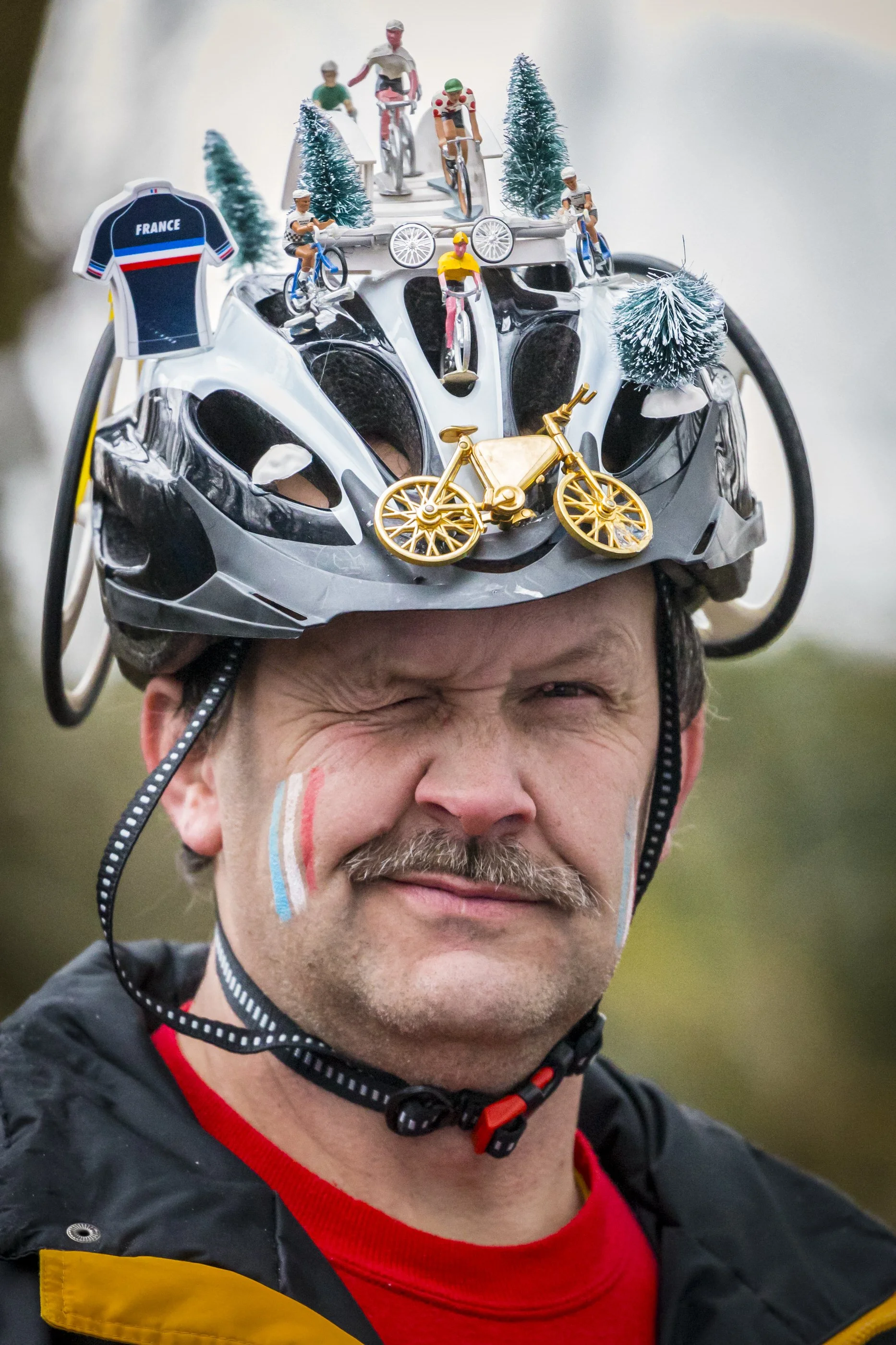 A man wearing a bicycle helmet decorated with miniature figures of cyclists, trees, and a France sports jersey, smiling with painted stripes on his face.