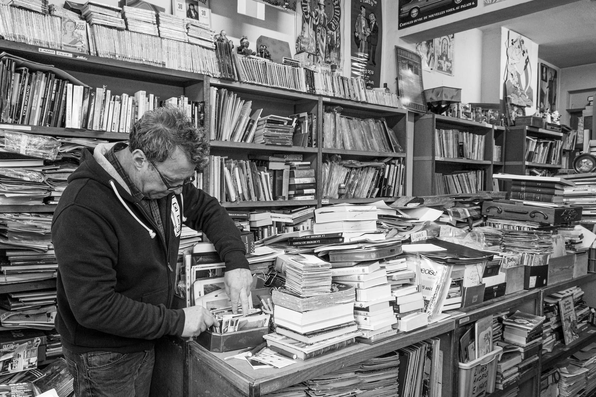 A man wearing glasses and a hoodie is sorting through books and papers on a table in a cluttered bookstore or library.