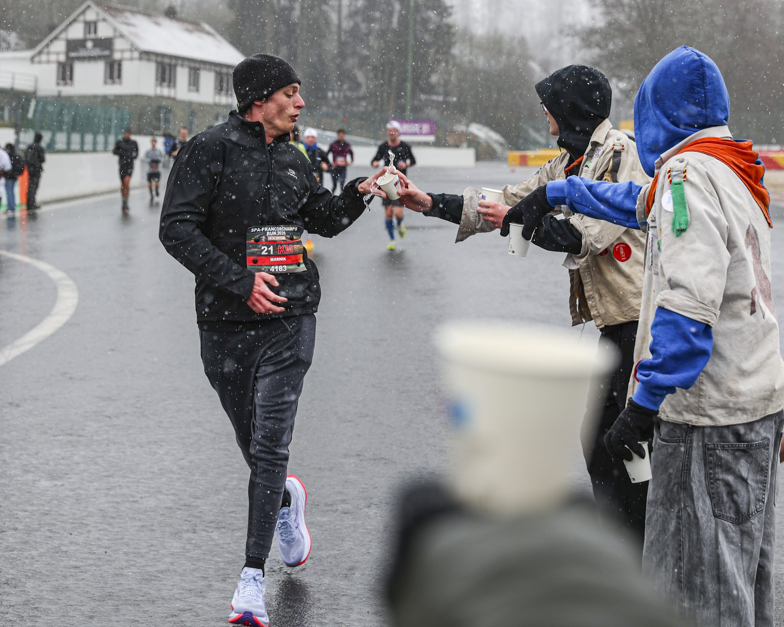 A runner in a black jacket and black beanie running in a rainy race, receiving a cup of water from a volunteer in a jacket and hood, with other runners in the background on a wet road