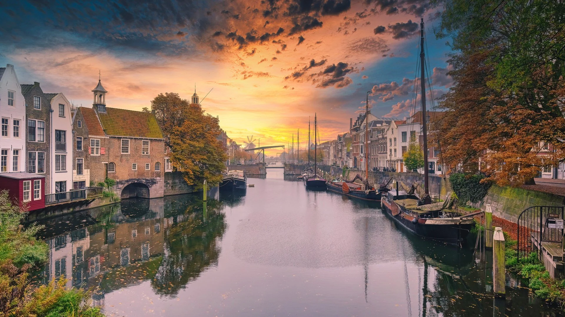 Sunset view of a canal with houses and boats, trees with autumn foliage, and a colorful sky.