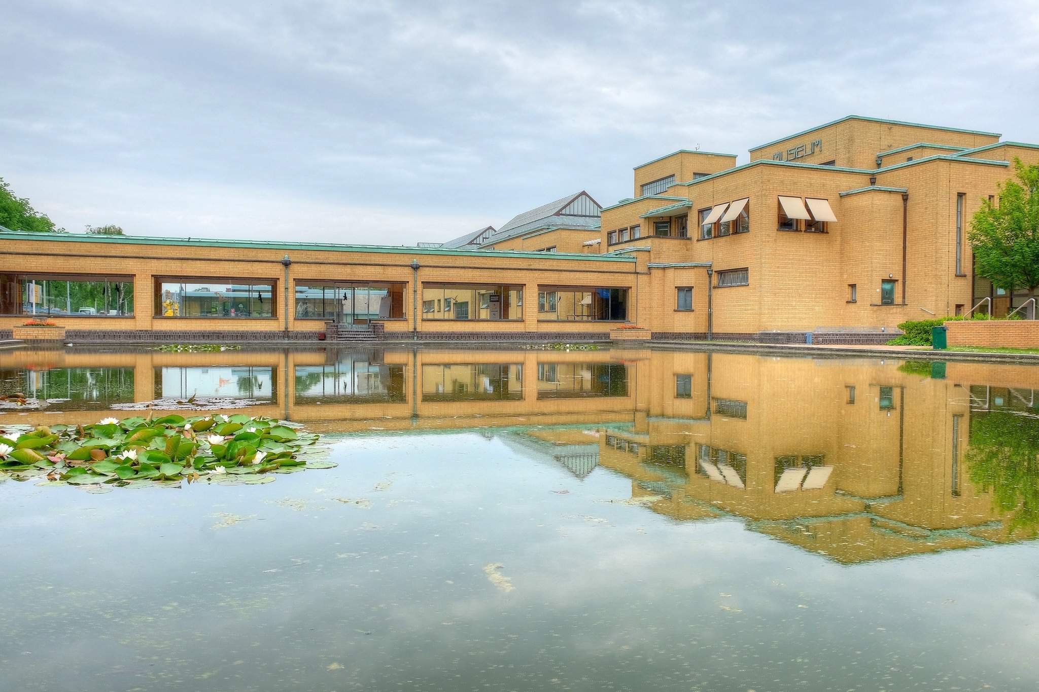 A modern brick building with large windows and a pond in the foreground, reflecting the building and cloudy sky.