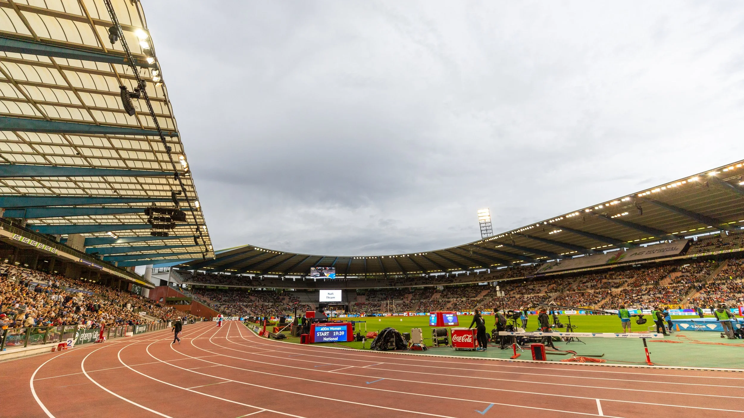 An outdoor stadium with a running track, a large digital screen at the center, and a crowd of spectators in the stands. The sky is overcast, and there are lights illuminating parts of the stadium.