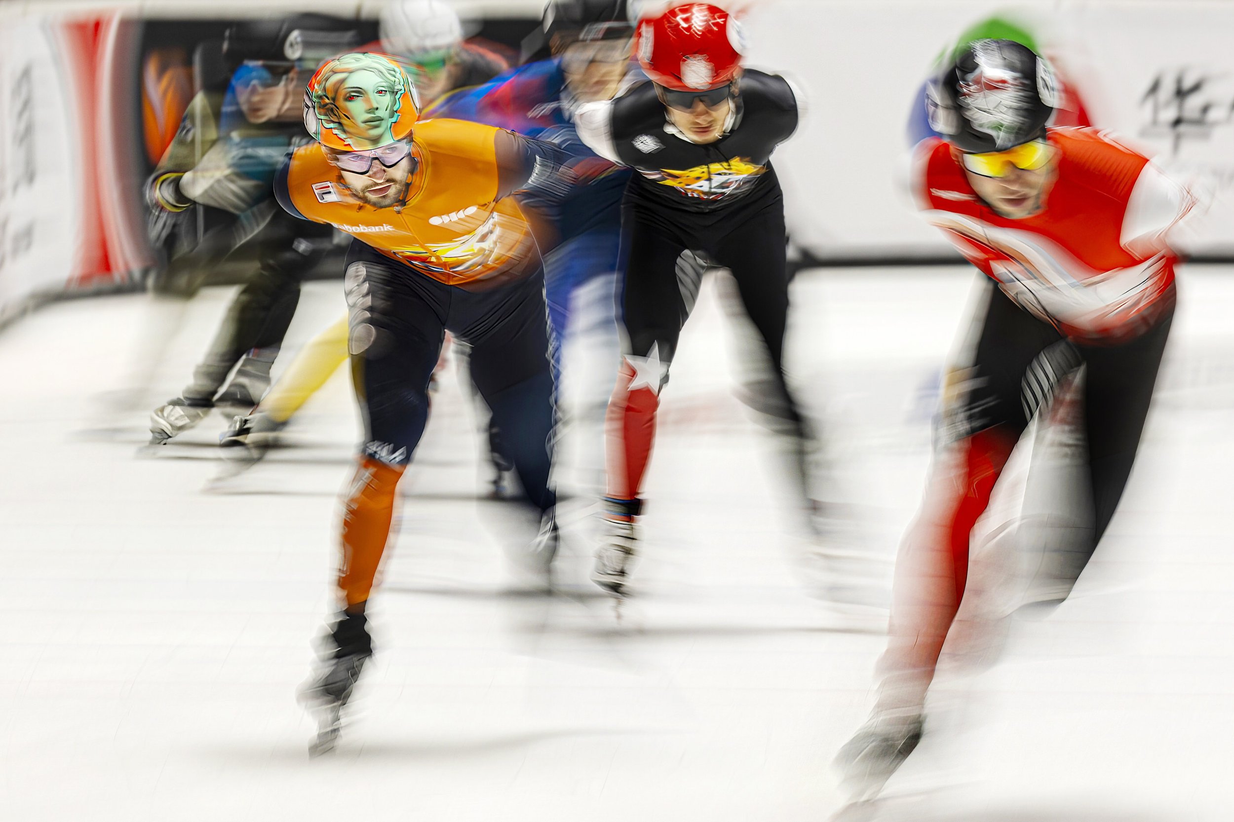 Four speed skaters in colorful uniforms and helmets race on an indoor ice rink, with a blurred background indicating fast motion.