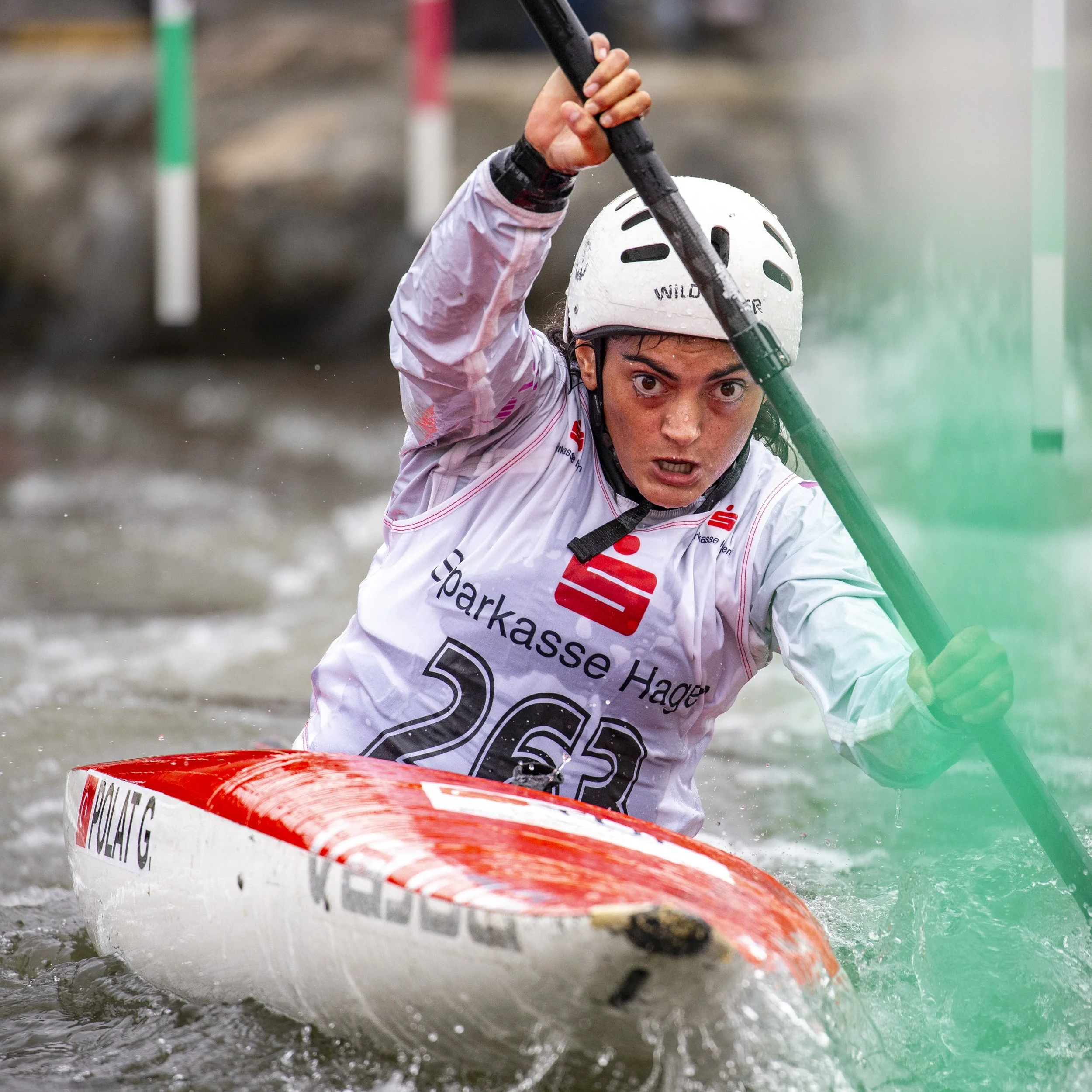 A person wearing a white helmet and a white jacket paddling a kayak through choppy water, holding a paddle high above their head, with a focused expression on their face.