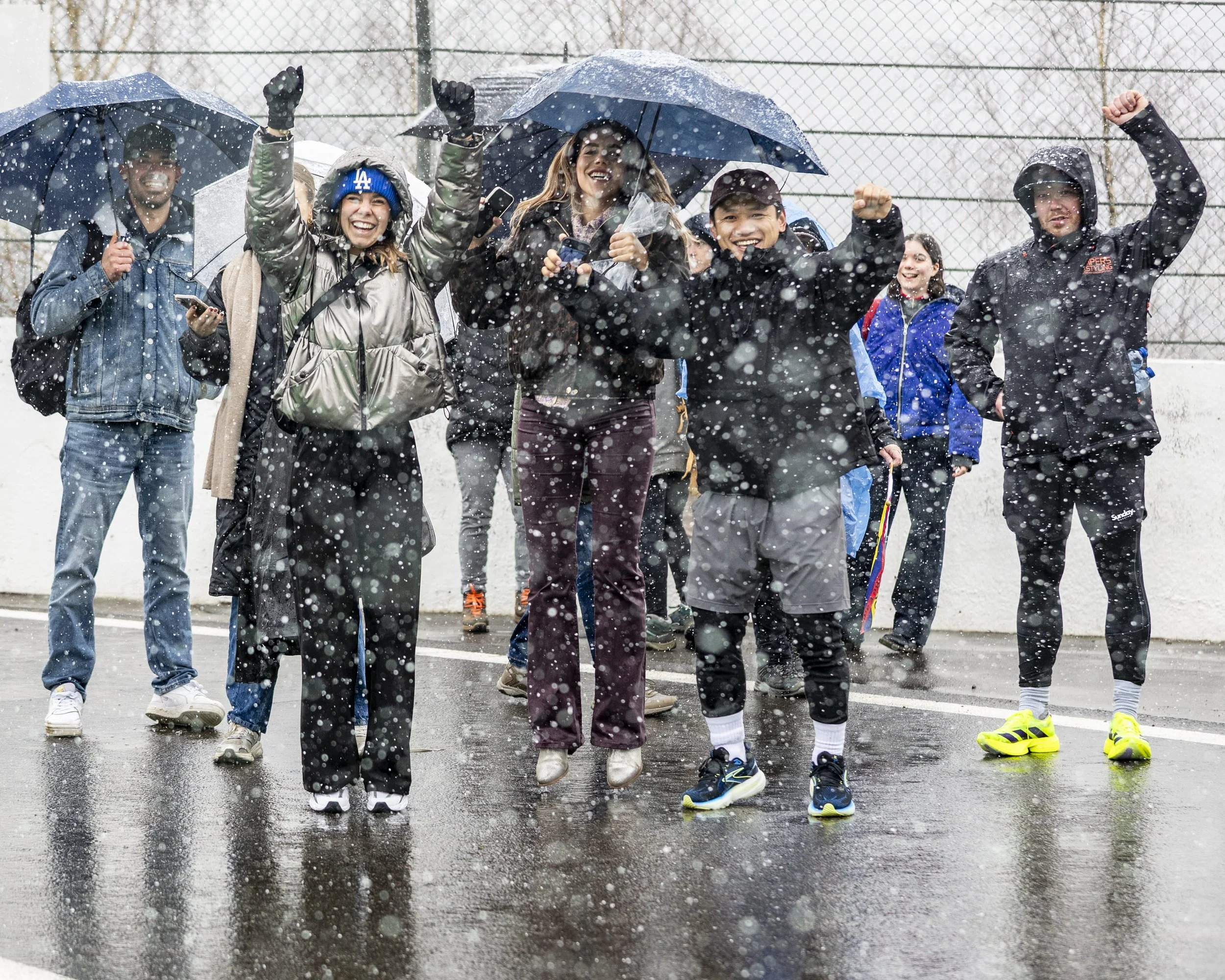 Group of people celebrating outdoors in rainy weather with umbrellas, jumping and smiling, on a wet paved surface near a chain-link fence.