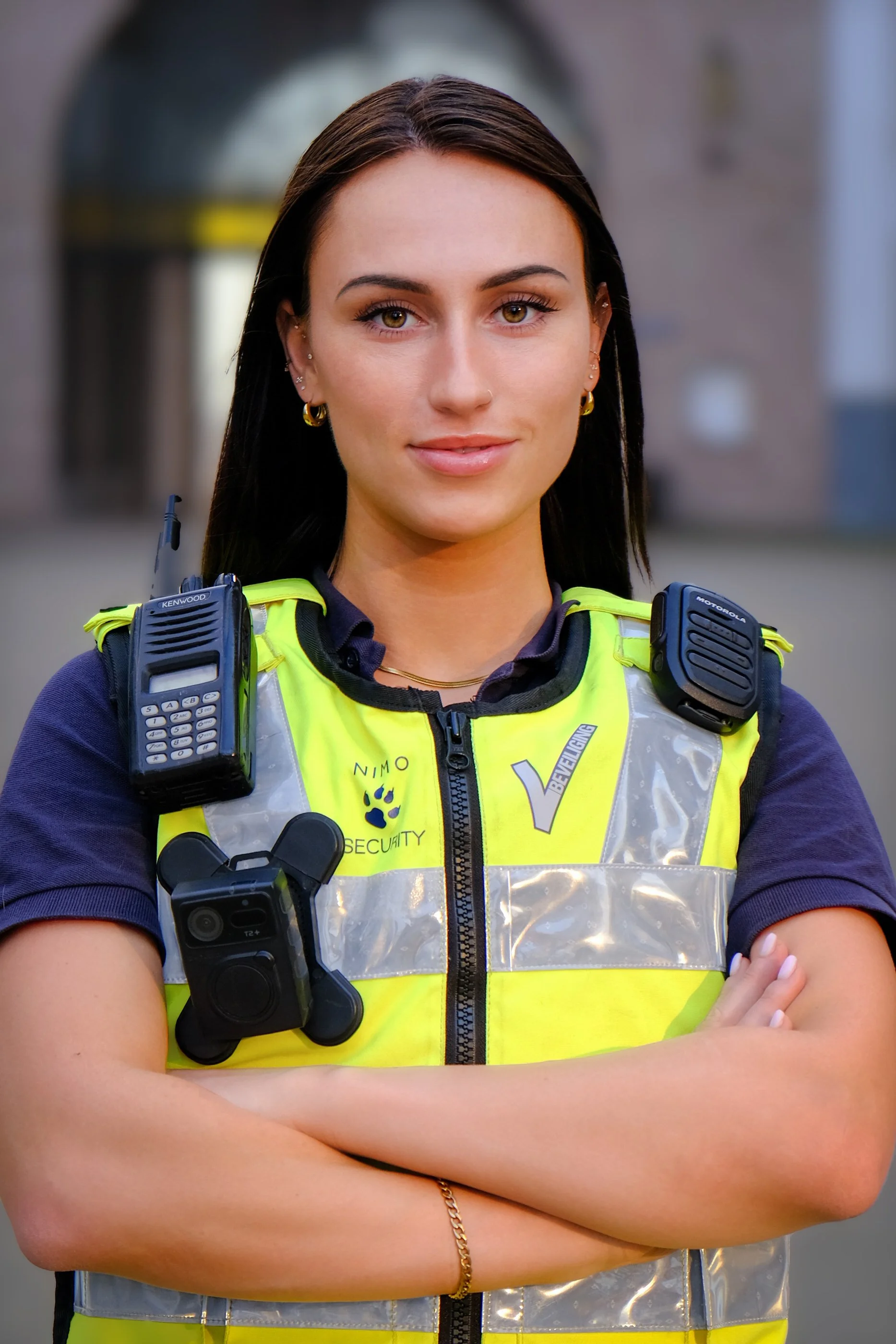 Young female security officer with dark hair, wearing a yellow reflective vest, standing outdoors with arms crossed, with various radios and communication equipment attached to her vest.