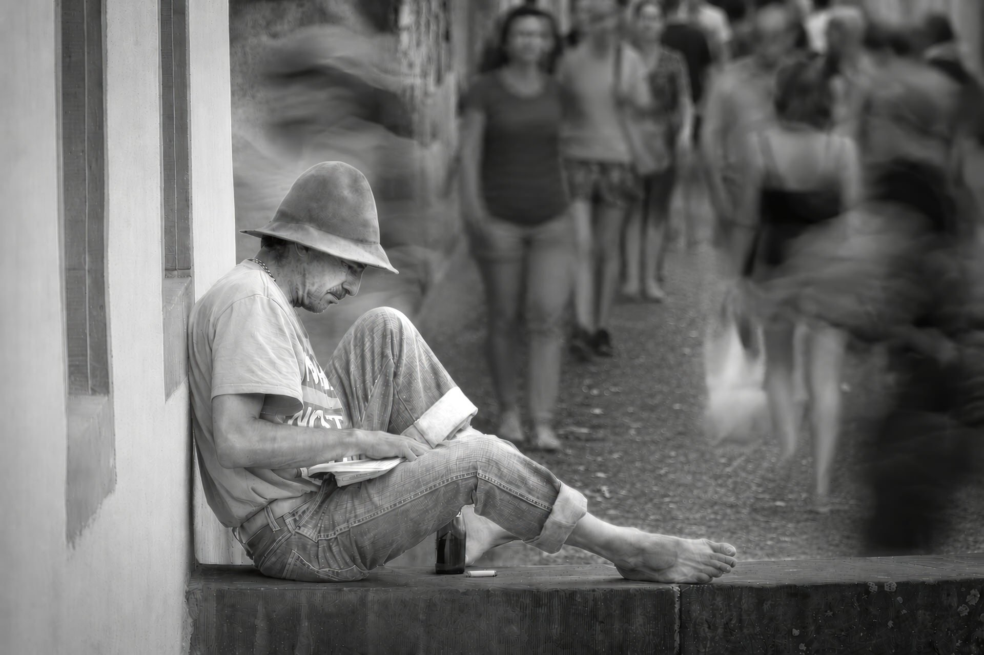 A man sitting on a ledge, wearing a helmet and casual clothes, reading a newspaper, with several blurred people walking in the background.