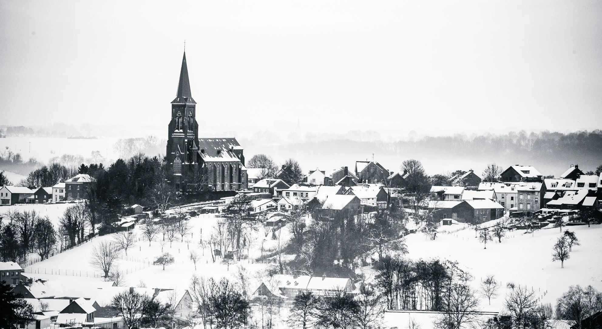A snowy village landscape featuring a large church with a tall, pointed steeple, surrounded by houses and trees blanketed in snow, with distant hills foggy in the background.