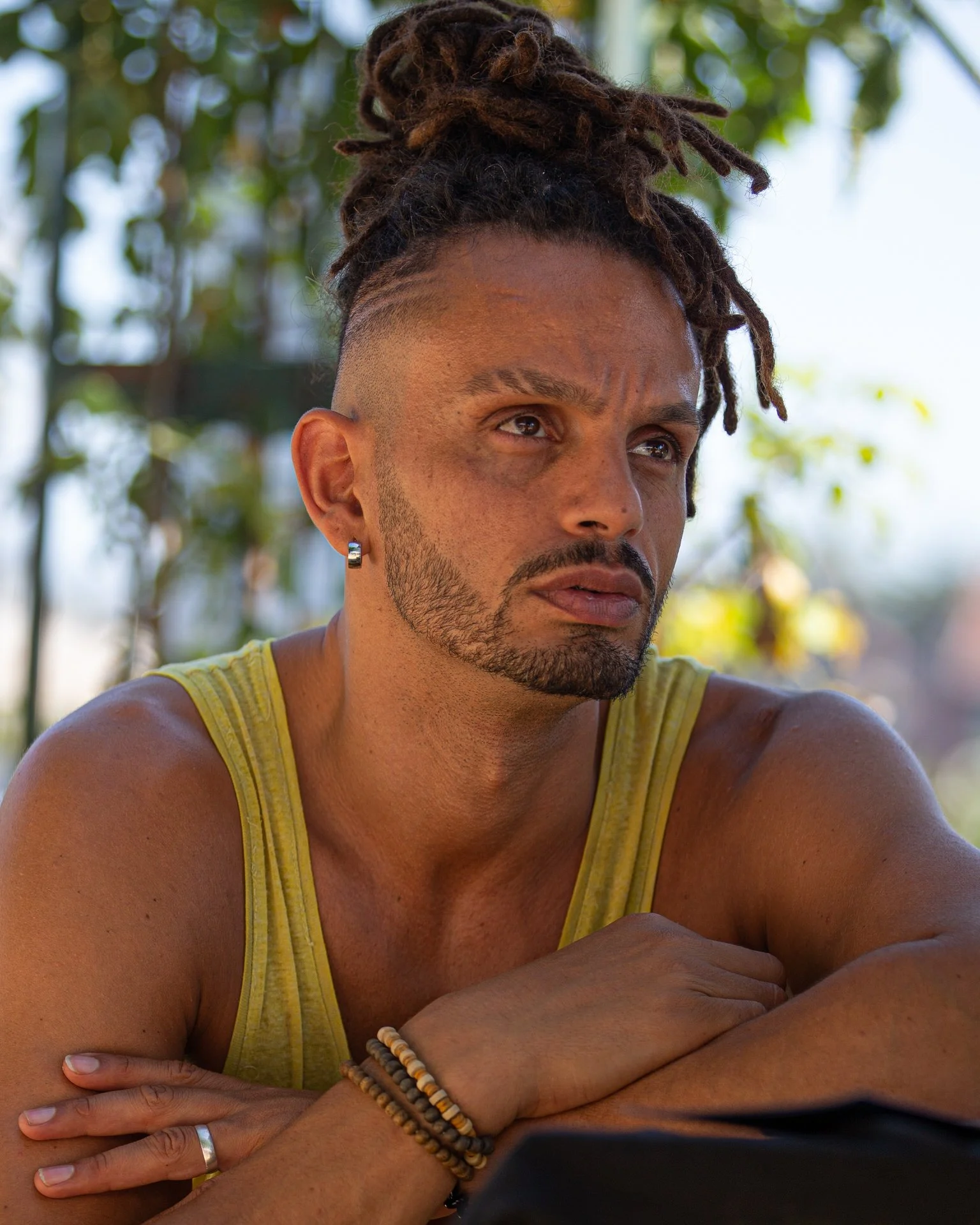 Close-up of a man with dreadlocks wearing a yellow tank top, earrings, and bracelets, sitting outdoors with a background of trees and greenery, looking serious and contemplative.